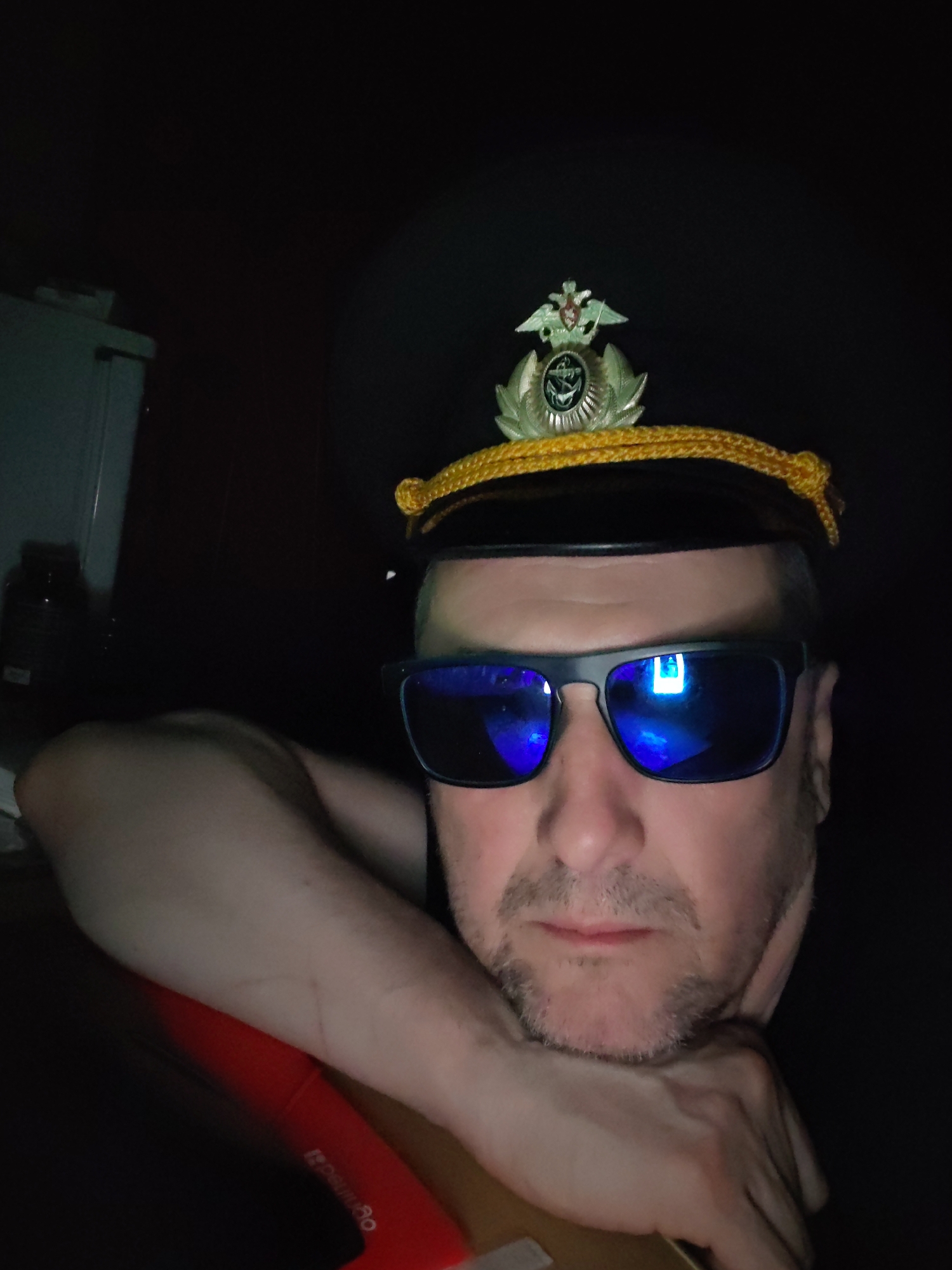 The image captures a dimly lit, close-up selfie of a man, likely taken indoors in Shcheleiki, Russia. The man, who appears middle-aged with short stubble, is wearing dark sunglasses with reflective blue lenses. He sports a dark cap adorned with a prominent gold-colored emblem featuring a two-headed eagle perched atop a wreath encompassing an anchor, indicative of a Russian Navy or maritime affiliation. A braided yellow cord wraps around the cap above the brim.
His right arm is bent, and his head rests on his hand, which is supported by a red object. This red object has partially visible white text that appears to be "ogitechlo," possibly part of a "Logitech" brand name. The background is mostly dark, with faint outlines of a dark bottle or container on the far left and a possible white appliance. A clear reflection of a bright, possibly rectangular light source, such as a computer screen, is visible in the right lens of his sunglasses. The overall darkness suggests the photo was taken at night or in a room with minimal lighting. The man looks directly at the camera with a neutral expression.