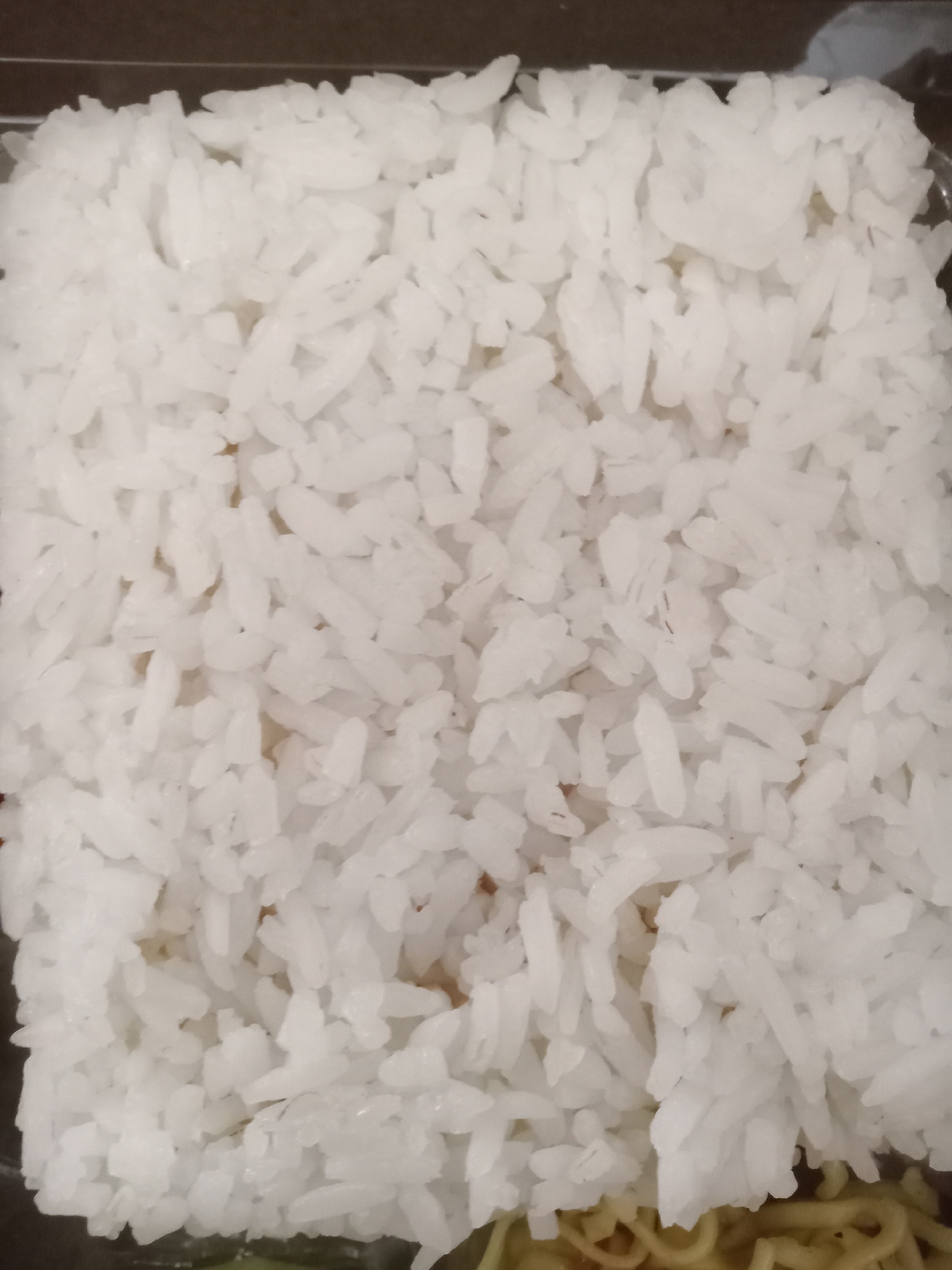 A close-up, overhead shot shows a rectangular portion of white rice filling most of the frame. The rice is slightly clumped in some areas, with individual grains visible. A dark, blurred background suggests the rice is contained in a box or container, with a hint of yellow noodles and green vegetables visible at the very bottom of the frame. The lighting is even, with no harsh shadows. There are no people or specific identifiable landmarks in the image to place it in Simo, Indonesia.