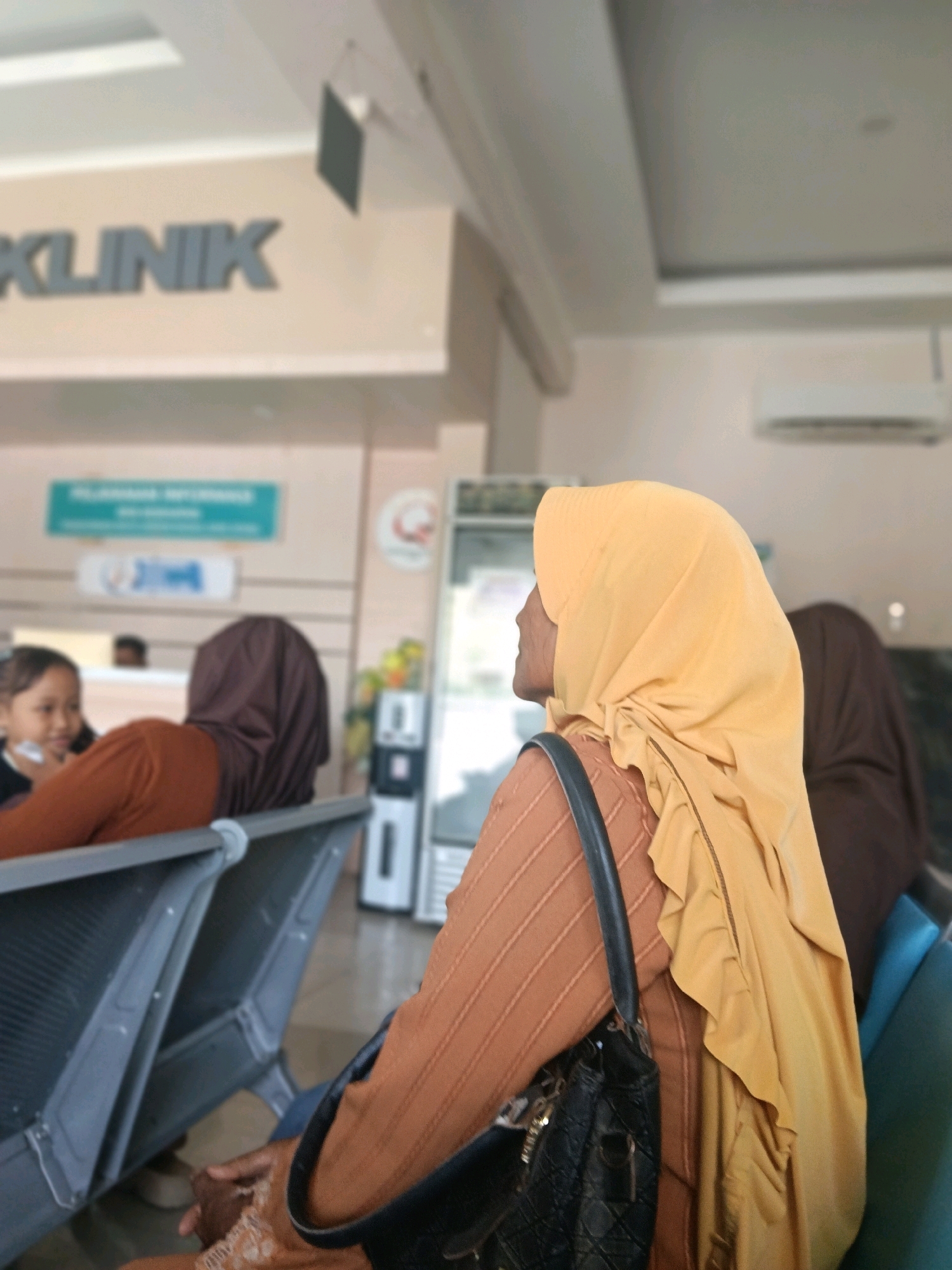 The image depicts the waiting area of a clinic, possibly in Balun, Indonesia. In the foreground, a woman wearing a yellow hijab and a brown, ribbed shirt is seated. She is looking to her left, with a black handbag slung over her shoulder. Her hands are visible in her lap, and she appears to be wearing intricate henna designs on her right hand. Behind her, another person wearing a brown hijab is seated, also facing away from the camera. To the left of the frame, a young girl with dark hair is visible, facing the person in the brown hijab and seemingly engaged in conversation. A man's head is partially visible in the background, near what appears to be a reception desk. The waiting area has gray plastic chairs arranged in rows. In the background, a sign with Indonesian text is mounted on the wall, and a sign with the word "KLINIK" is also visible. Other elements in the environment include what appear to be air conditioning units mounted on the ceiling and a water dispenser. The lighting suggests an indoor setting with overhead illumination.