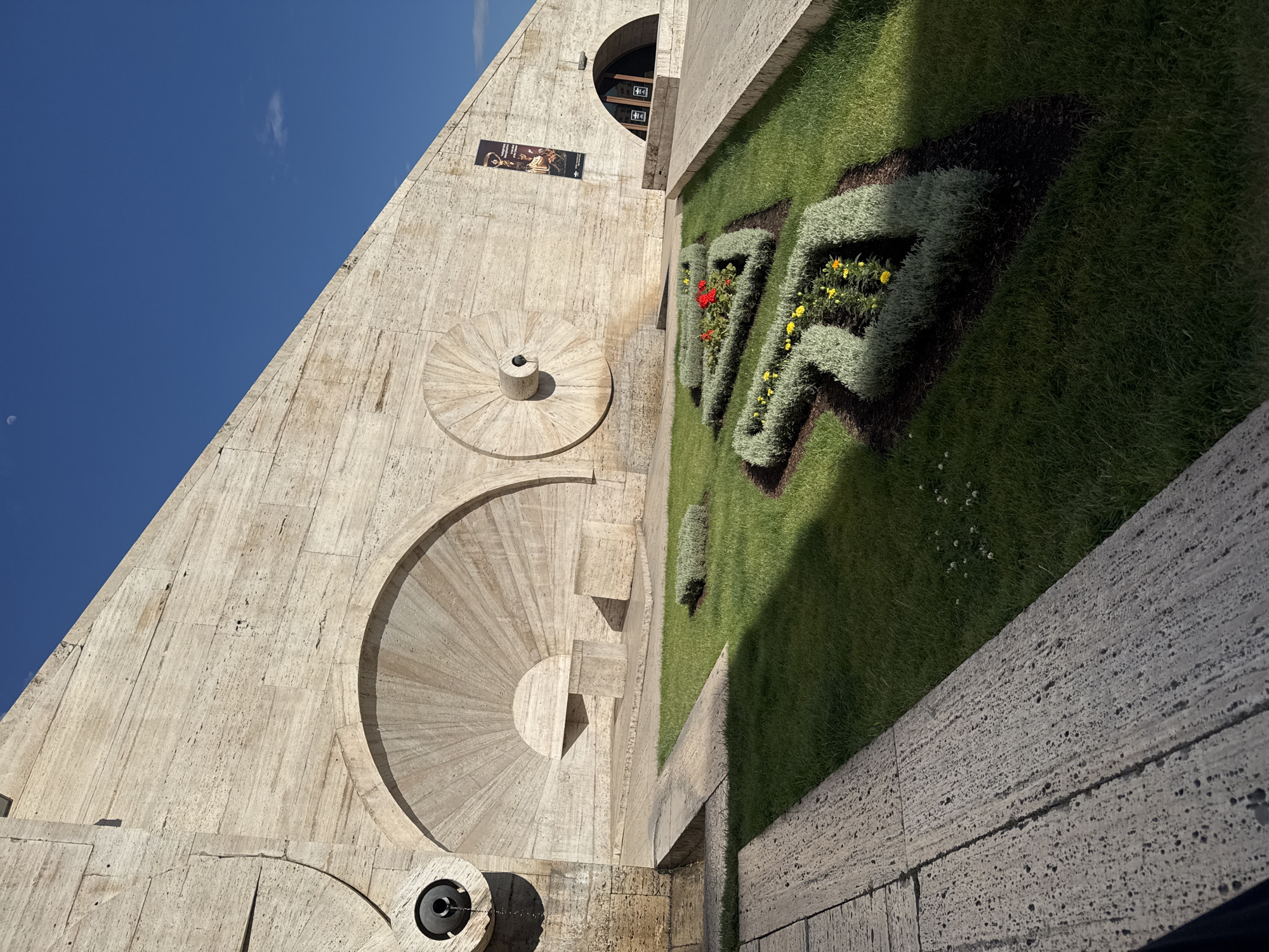 This image depicts a partial view of the Cascade in Yerevan, Armenia. The Cascade is a monumental stairway with fountains and sculptures, linking the downtown Kentron area of Yerevan with the Monument neighborhood. The image showcases the distinctive white travertine stone architecture of the structure, featuring circular fountains and sculpted elements. A poster is visible on the wall near an arched opening. Adjacent to the Cascade's structure is a meticulously manicured garden with geometrically shaped shrubs and flowers. The sky is clear and blue with a faint moon, indicating it is daytime. The image is taken at an angle, emphasizing the upward slope of the Cascade.