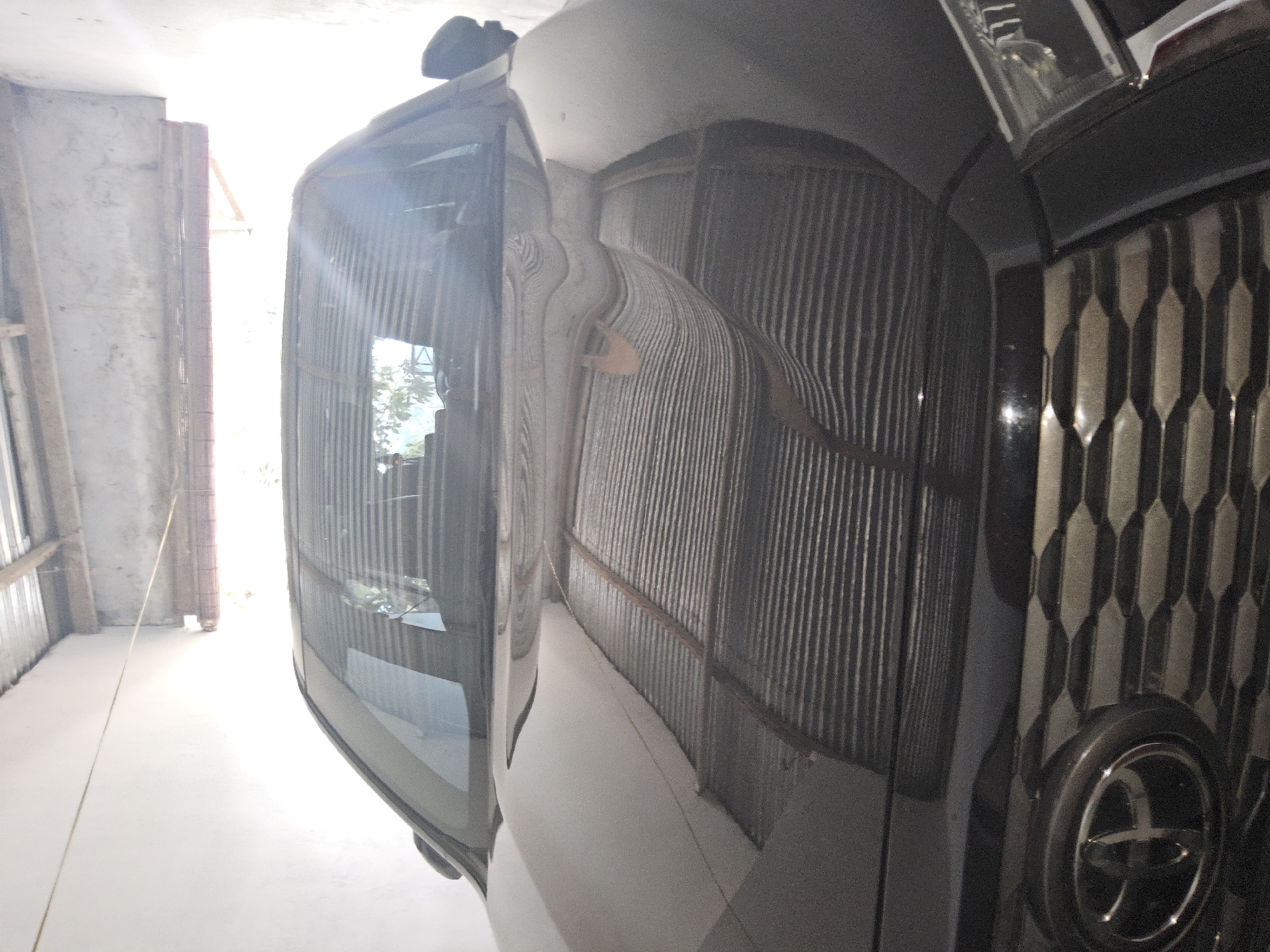 A black Toyota SUV is parked inside a covered garage. The garage has a corrugated metal roof supported by wooden beams, and the walls appear to be white plaster. The car's hood and windshield reflect the corrugated roof, creating distorted patterns. The front grille of the car, featuring the Toyota logo, is visible. The environment suggests a warm climate, possibly residential. The location is confirmed to be in Payagaboh, Indonesia.