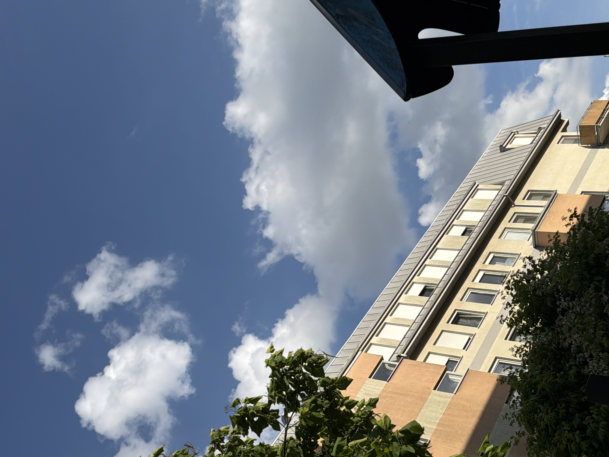 The image depicts a view of a building against a sky with clouds. The building appears to be multi-story with an orange and yellow facade. The roof is a grey, sloped metal structure. There are several windows on the building, some with closed shutters. Trees with green leaves are visible in the foreground, partially obscuring the lower portion of the building. In the right foreground, a dark, curved object, possibly part of a structure or awning, juts into the frame. The sky is blue with scattered white cumulus clouds. The lighting suggests it is daytime, with sunlight casting shadows. The location is identified as Unknown, Unknown.