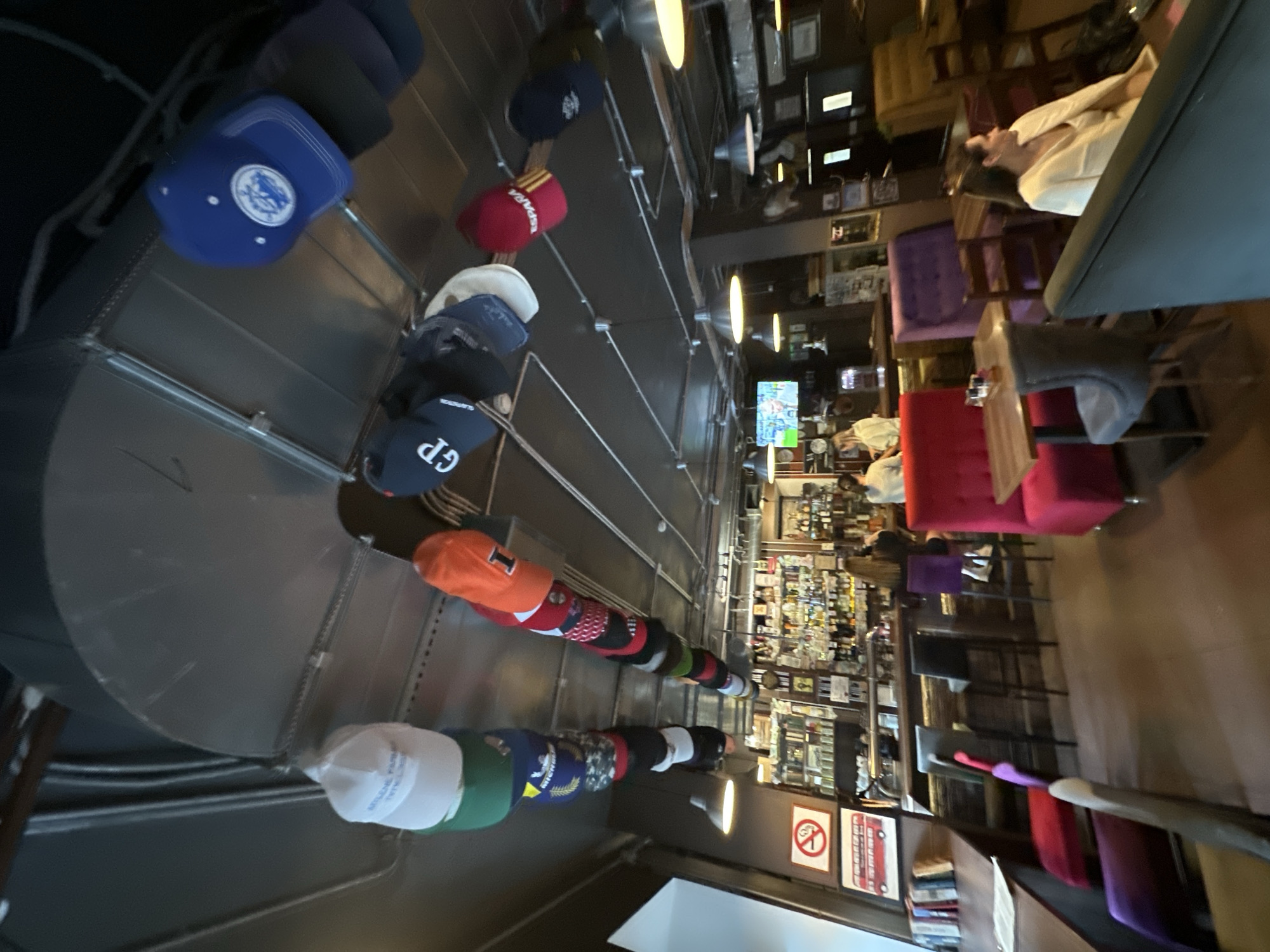 This image depicts the interior of a bar or restaurant located in Moscow, Russia. The ceiling is characterized by exposed ductwork and industrial-style pendant lights, from which numerous baseball caps are suspended in two linear arrangements. Identifiable caps include a white cap with "Mylene Farmer" text, a navy blue cap with "GP" embroidered, an orange cap featuring the letter "I", a red cap displaying "ESPARA", and a blue cap with a white circular logo.
On the left wall, a standard "No Smoking" sign is displayed above a framed image of a red double-decker bus. Below these, a shelf holds a collection of books. The central feature is a well-stocked bar counter, displaying various bottles and glassware, with multiple bar stools featuring purple, grey, and black upholstery. One individual with long brown hair is seated at a purple bar stool with their back to the camera, facing the bar. Two additional, blurred individuals are visible behind the bar.
Seating arrangements include a red upholstered booth and table on the left, accompanied by a grey upholstered chair. Further back, a purple upholstered booth is visible with a wooden chair. On the right side of the frame, a woman with brown hair, wearing a light-colored garment, is seated at a table, facing towards the left. A television screen displaying a blurred male face is mounted above the bar area. The floor throughout the establishment appears to be a consistent reddish-brown.