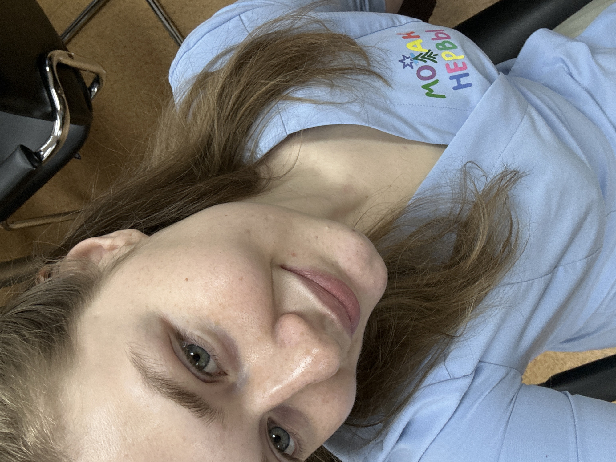 A person with light brown hair and gray eyes, wearing a light blue shirt with the Russian phrase "Мои нервы" (My nerves) embroidered in colorful letters, possibly a slogan or humorous expression, appears to be in an indoor environment. The person is sitting in a black office chair with a chrome frame, with a wooden floor visible in the background. Given the text on the shirt is in Russian, the location may be Kazan, Russia, but there is no specific detail to prove it.