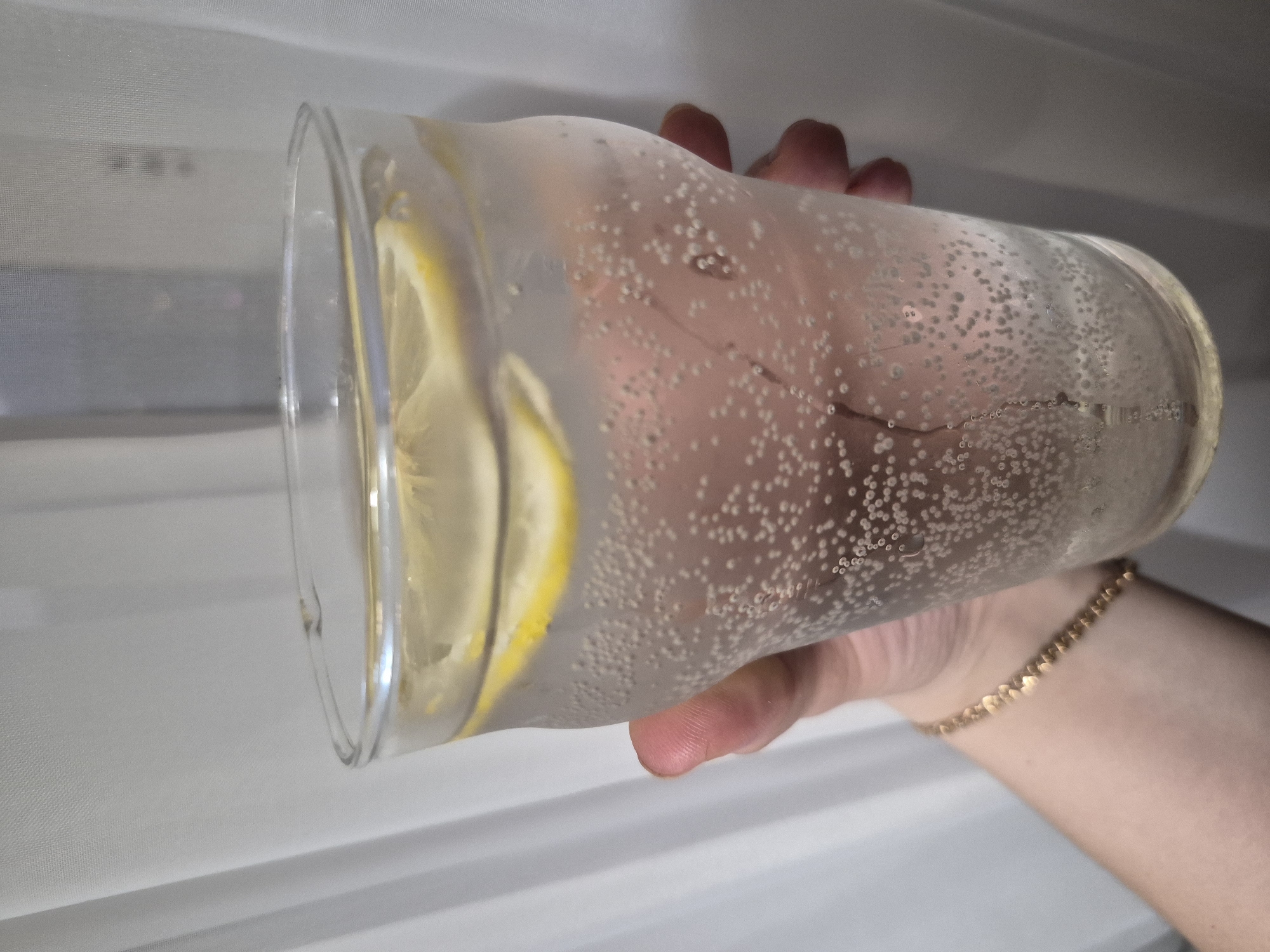 A hand holds a clear glass filled with a bubbly liquid and lemon slices. The bubbles suggest the drink is carbonated, possibly sparkling water or a similar beverage. The background features a white, possibly fabric, surface with horizontal lines, resembling curtains or blinds. The composition focuses on the hand and glass, with the background blurred.
The image doesn't provide any information to infer the location is Tobolsk, Russia.