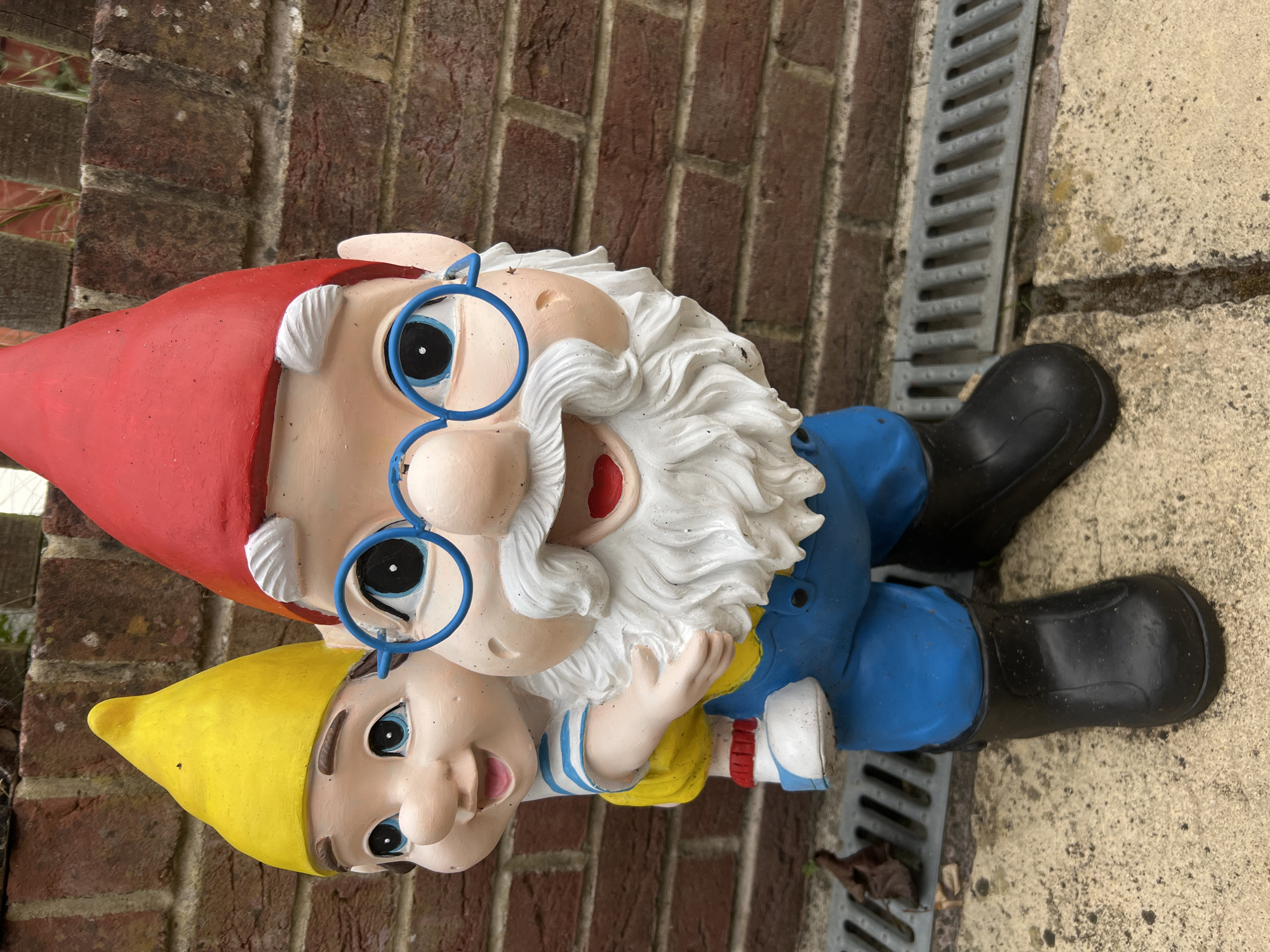A garden gnome statue of two gnomes is leaning against a brick wall. The top gnome has a red hat, white beard, and blue glasses. The bottom gnome has a yellow hat and a smiling face. The statues are located in Basingstoke and Deane, United Kingdom. Next to the gnome there is a grey gutter.