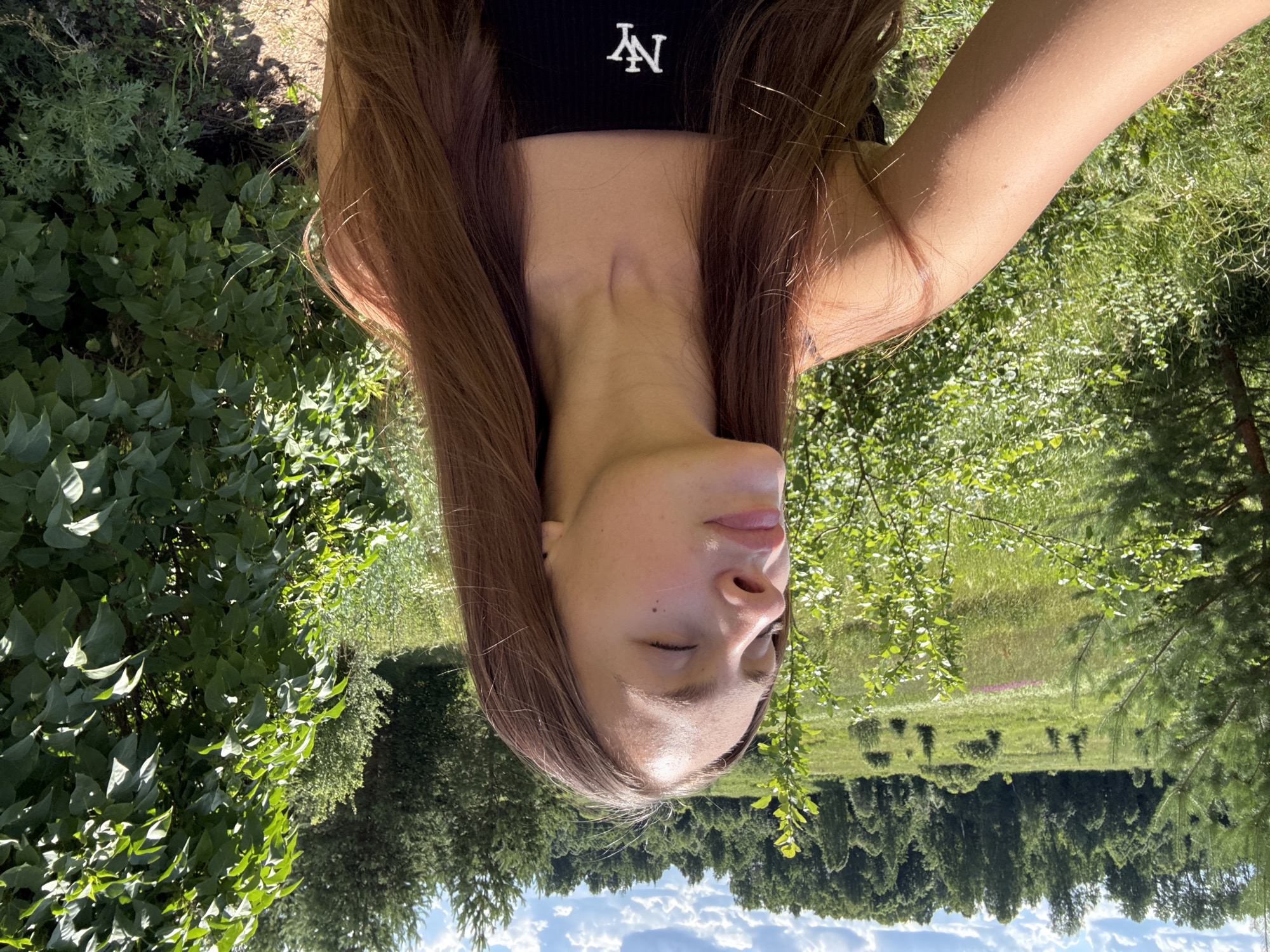 This is a portrait of a young woman taken outdoors. She has long, brown hair that frames her face. Her eyes are closed, and her head is tilted back slightly. She is wearing a black tank top with a white "NY" logo on the chest. The background is a lush, green landscape with trees and grass, suggesting a natural setting. The lighting indicates it is daytime, likely sunny, given the bright illumination. The image appears to be taken in Шолохово, Russia, as indicated by the location context. The overall mood is serene and relaxed, with the subject enjoying the outdoors. The composition of the photo, with the subject positioned prominently against the natural backdrop, emphasizes her presence within the environment. The foliage is vibrant green, indicating it is likely summer or late spring. The sky is visible with some clouds, further confirming it is daytime.