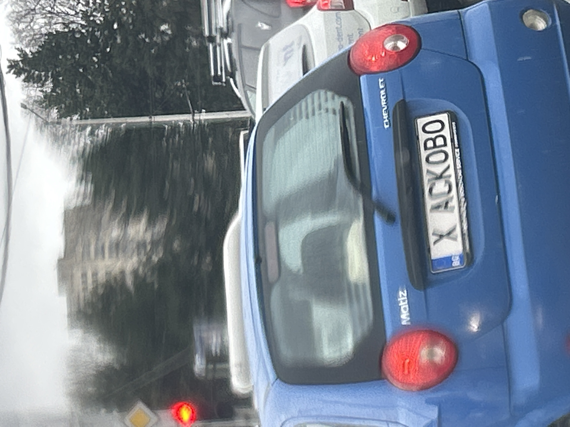 The image shows the rear of a blue Chevrolet Matiz car with a Bulgarian license plate "X ACK0BO" in Sofia, Bulgaria. The car is stopped in traffic. Behind it, another vehicle is partially visible. In the background, a blurred cityscape and trees are visible through a wet windshield. A red traffic light is also present.