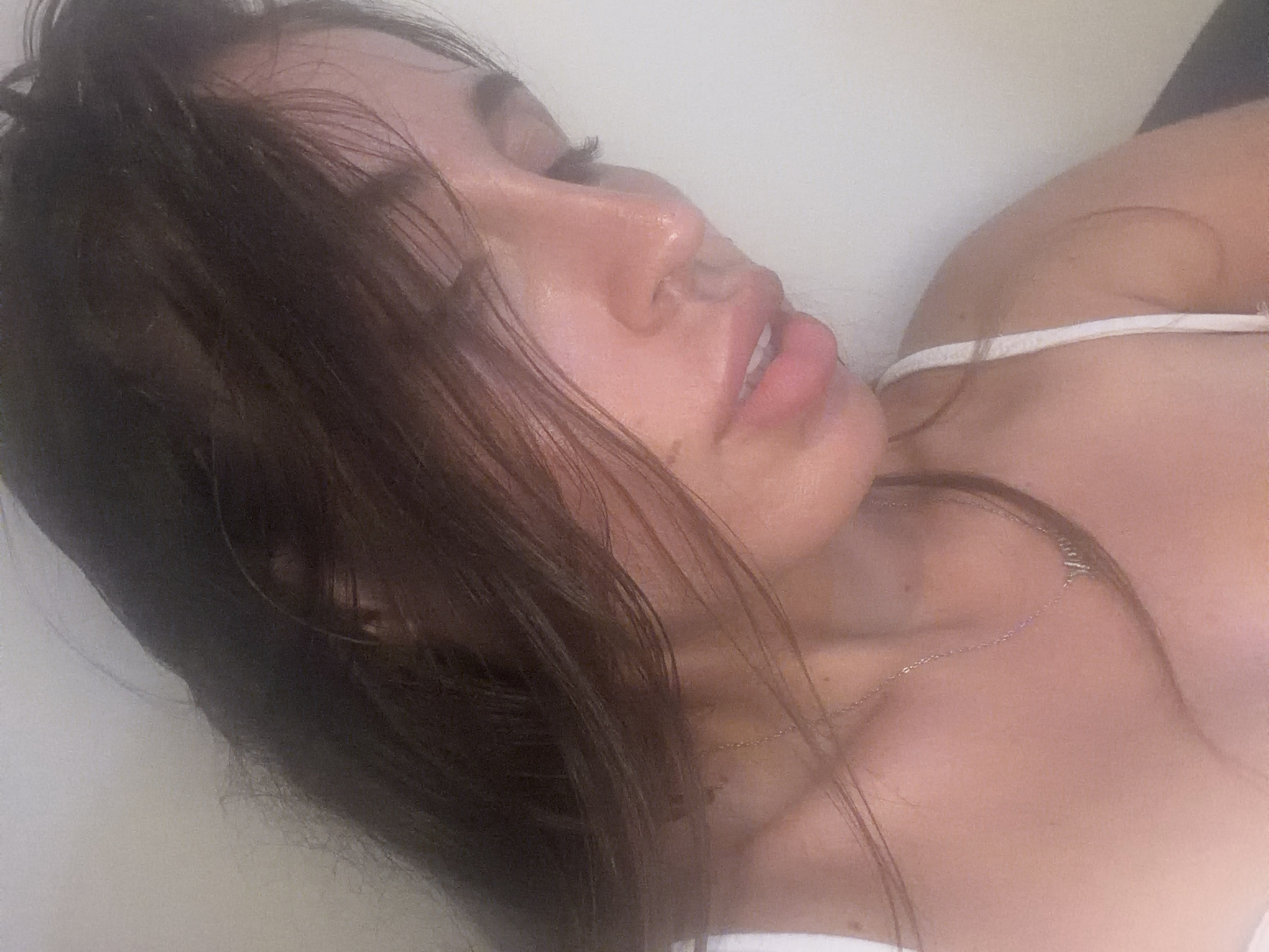 A close-up, waist-up, over-the-shoulder shot captures a young woman with long, dark hair against a plain, light-colored wall. Her hair is partially covering her face, obscuring one eye and the left side of her forehead. She is wearing a light-colored tank top with a thin strap visible on her right shoulder. Around her neck, she wears a delicate silver chain with a pendant. The lighting is soft and diffused, creating a slightly hazy effect on her skin, which appears to have a dewy sheen. Her mouth is slightly open, and her gaze is directed downwards and to the left. The scene is intimate and personal, with no discernible background details suggesting a specific location within Tersefanou, Cyprus, or any other city. The focus is entirely on the woman and her immediate features.