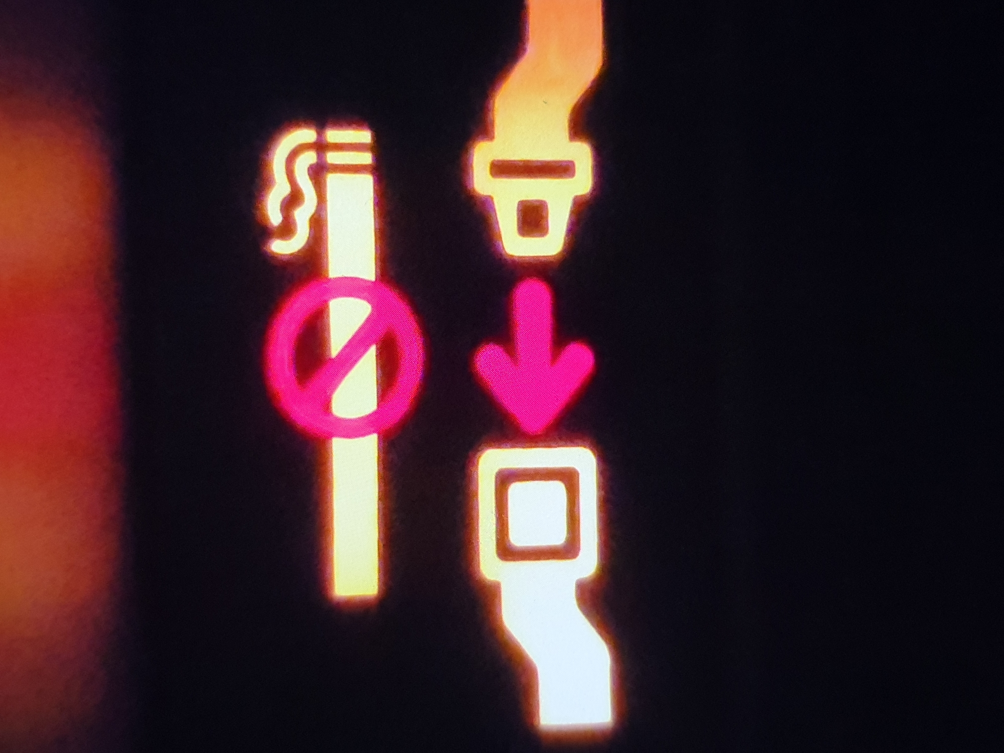 The image shows two illuminated symbols on a dark background. On the left is a no-smoking symbol, with a cigarette that has smoke coming from it, and a red circle with a slash across it. On the right is a seatbelt symbol, with an arrow pointing to the seatbelt buckle.