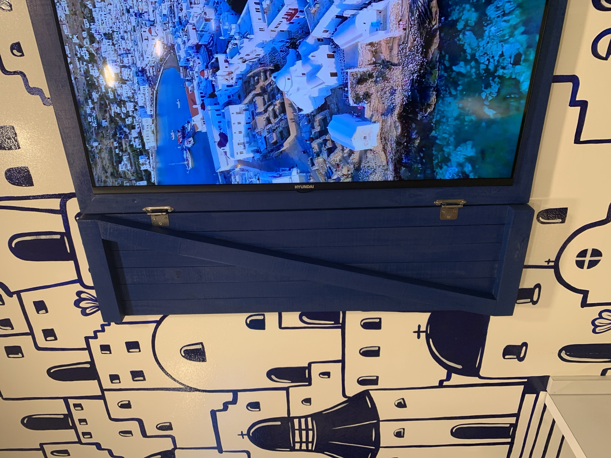 An indoor scene captures a wall-mounted television and a decorative wooden shutter below it. The television displays a vibrant aerial view of a coastal town, characterized by numerous white-washed buildings nestled along a rocky coastline. A clear blue bay with boats docked in a harbor is prominent, and the water transitions to a lighter turquoise near the shore, suggesting shallowness. The scene on the screen is indicative of a bright, sunny day in a Mediterranean or Greek island setting. The word "HYUNDAI" is visible on the bottom frame of the television.
Below the screen, a dark blue, wooden shutter, constructed from planks and featuring a diagonal brace, is closed. Two silver-colored metallic latches are secured on its top edge. The wall surrounding both the TV and the shutter is painted white and adorned with a stylized mural. This mural depicts outlines of buildings, bell towers, and arched windows in dark blue, complementing the screen's imagery and creating a cohesive Mediterranean village theme. Although the provided context is Pampatar, Venezuela, the visual elements, especially the TV content and wall mural, evoke the architecture and scenery typically found in Greek islands.
