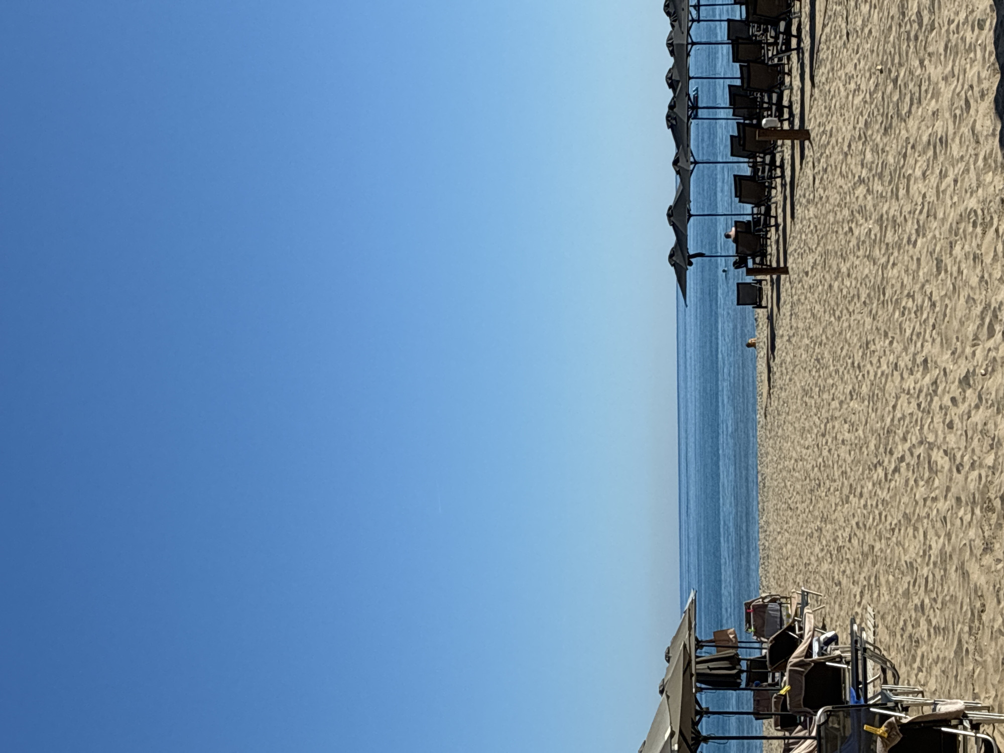 A bright, sunny day at a beach in Rethymno, Greece. The sand is light brown and textured. In the foreground, lounge chairs and umbrellas are neatly arranged on the sand. To the left, several light-colored umbrellas and lounge chairs are visible, some with towels draped over them. To the right, a row of dark umbrellas shades more lounge chairs. A few people are present; one person is sitting under an umbrella on the right side, facing the sea. Another person is visible in the water to the left, further out from the shore. The sea is a calm, deep blue, meeting a clear, bright blue sky at the horizon. The overall scene is tranquil and indicative of a typical beach day.