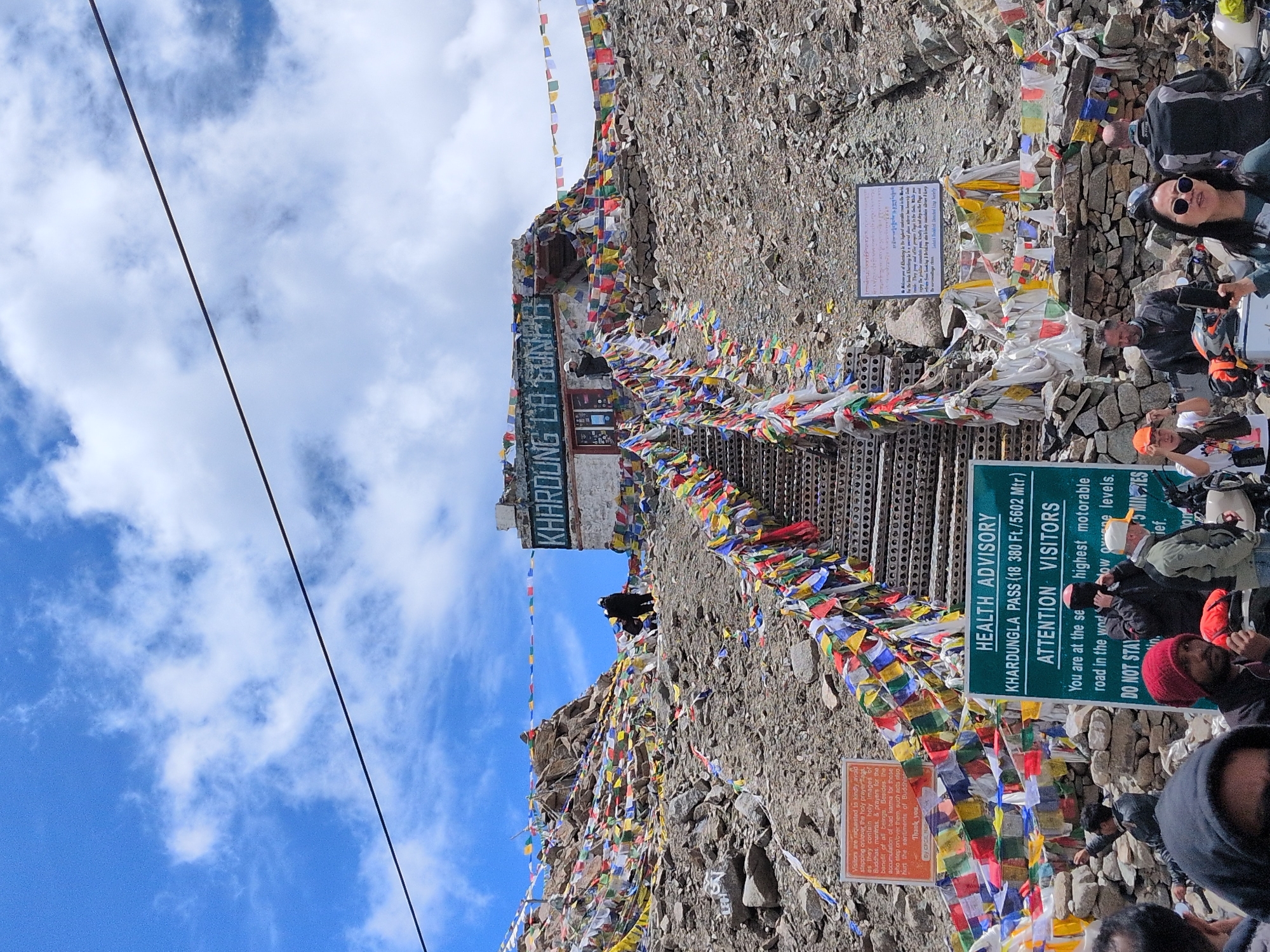 The image captures a view of the Khardung La Pass in Sasoma, India, a mountain pass known for being one of the highest motorable roads in the world. A steep set of metal stairs, adorned with numerous colorful prayer flags, ascends a rocky hillside towards a building at the summit. The building, with signage that reads "Khardung La," is also decorated with prayer flags. Several signs are visible at the base of the stairs, including one labeled "HEALTH ADVISORY" for Khardungla Pass, providing altitude information and advice for visitors. Another sign, with orange text on a white background, states "Visitors are requested to kindly avoid stepping onto the holy images..." A group of people, dressed in casual and outdoor wear, are gathered at the bottom of the stairs, some appearing to be tourists. A few individuals are seen interacting with each other, and some are holding cameras or phones. The environment is mountainous and rocky, with a clear blue sky filled with white clouds. A power line stretches across the top of the frame.