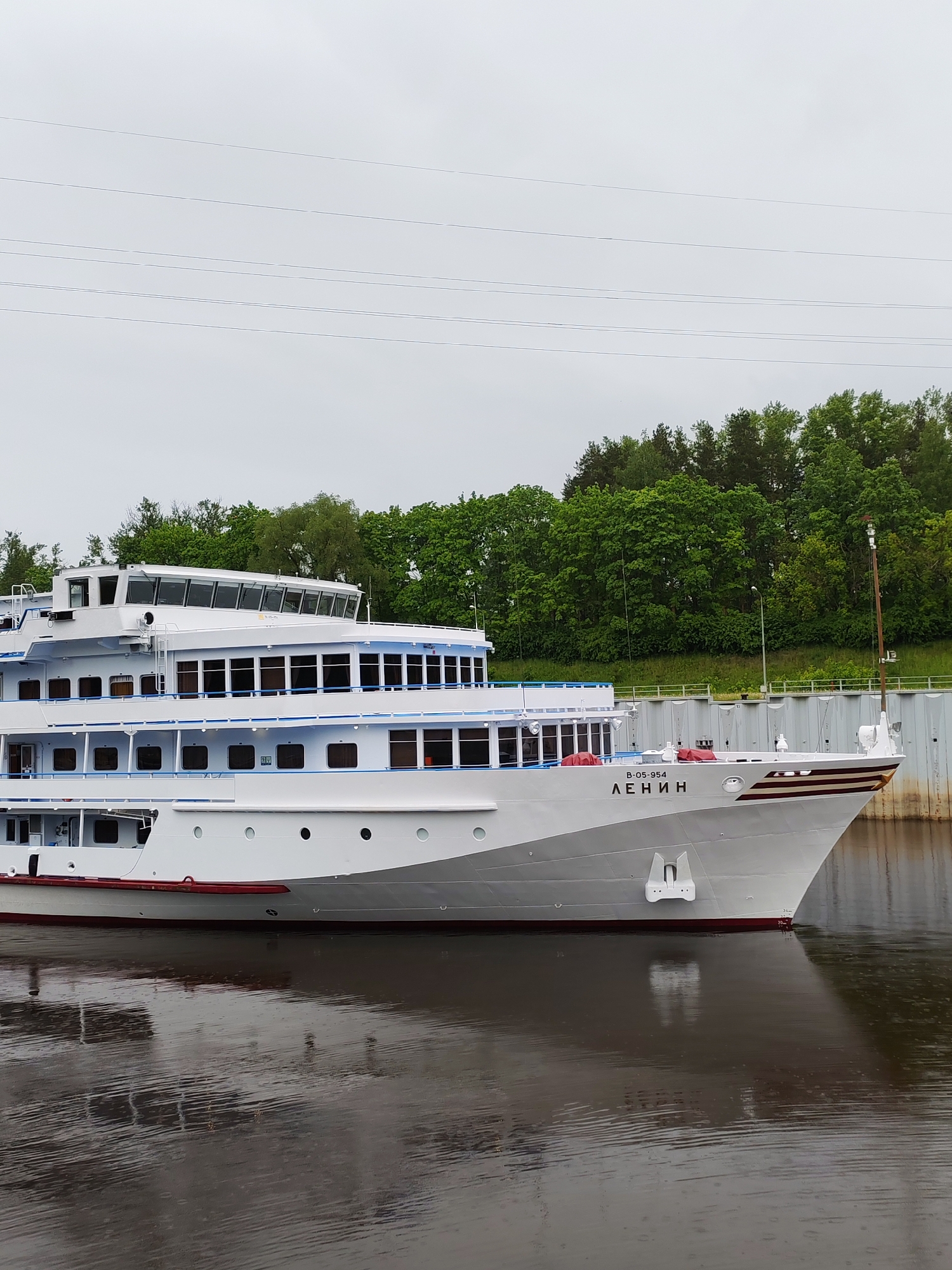 The image shows a white cruise ship named "Lenin" (written in Cyrillic) docked in a waterway. The ship has multiple decks, and a red stripe is visible along the hull. The surrounding environment features green trees on the bank and a concrete structure in the background. Overhead power lines are also visible against the overcast sky. The water is calm with visible ripples. The location is likely near Bol'shie Mkhi, Russia, given the ship's name and style.