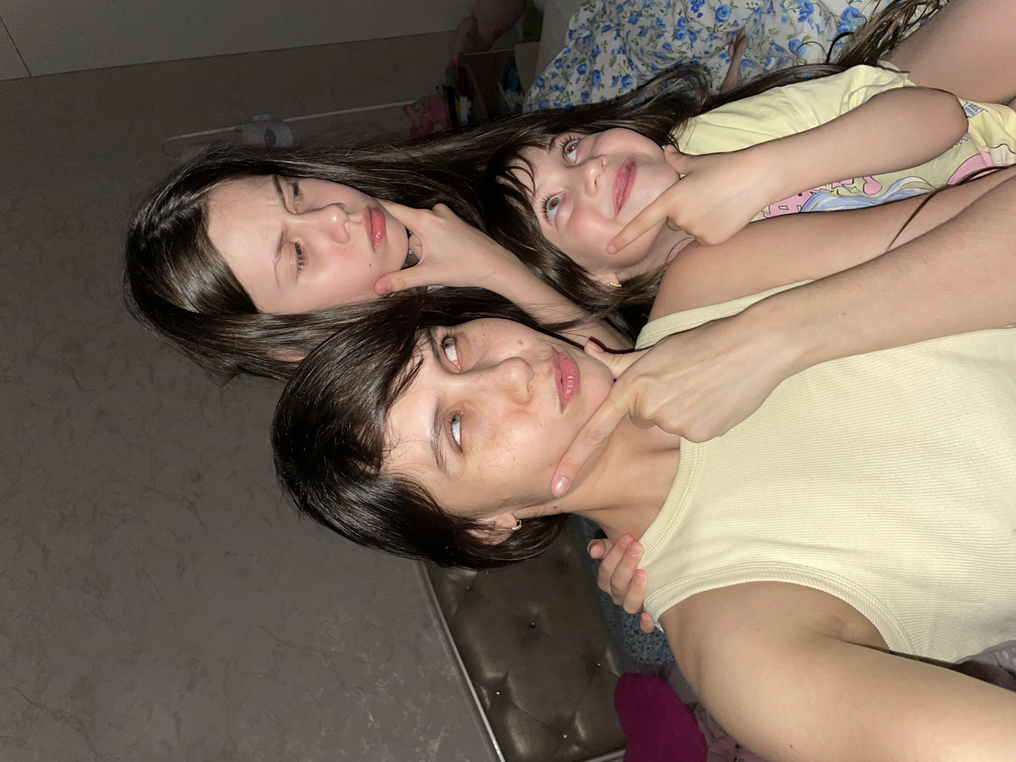 Three people are lying on a bed with a floral patterned bedspread. The person in the foreground is wearing a yellow top, and is lying on their back with their head angled slightly to the right. The middle person is lying on their back and has their left hand touching their chin. The third person is on their back and is holding their right hand to their chin. All three people have brown hair. The background is a light gray wall. The location cannot be verified as Izhevsk, Russia.