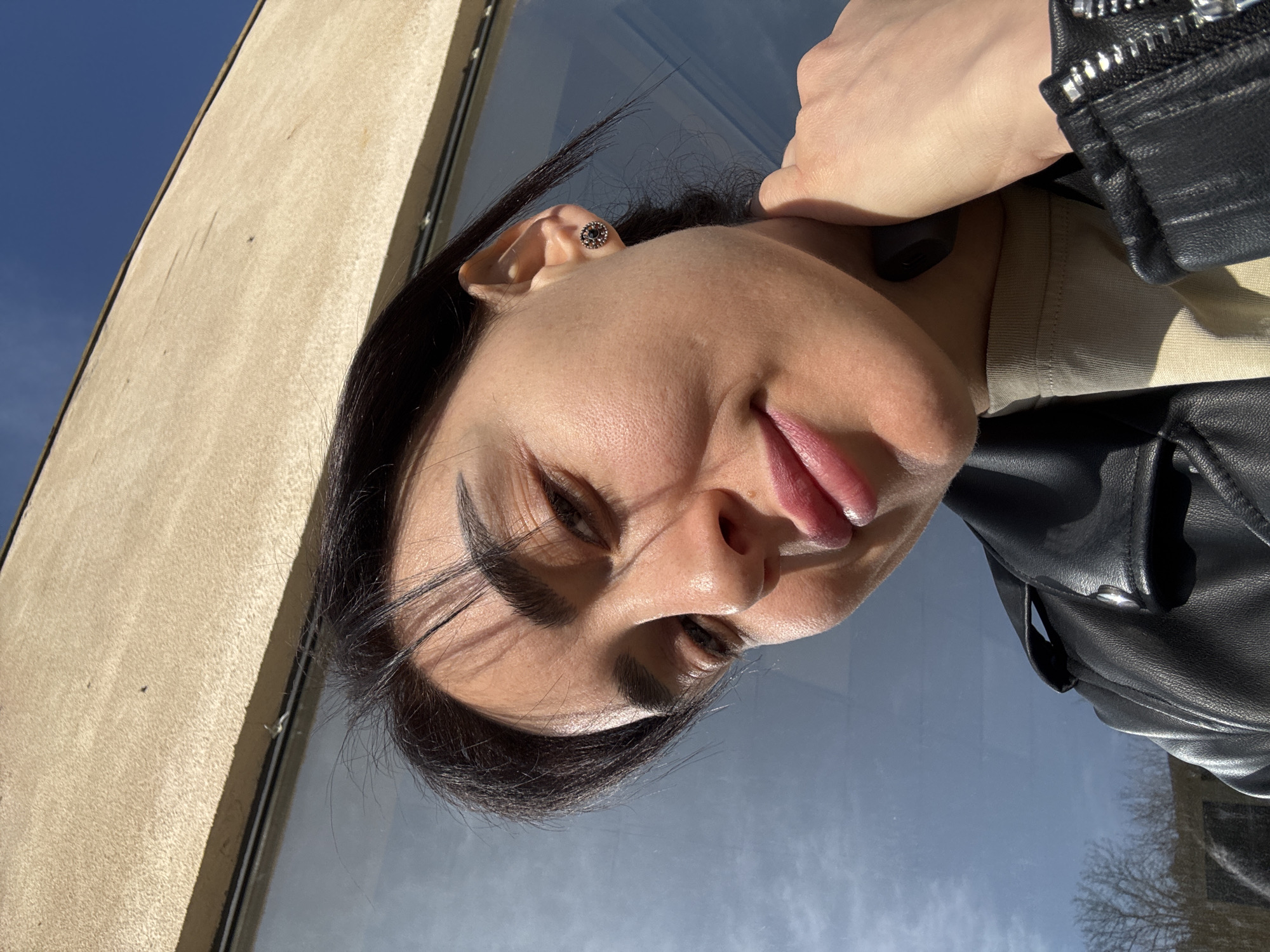 A close-up, upward-angled shot shows a woman with dark hair and fair skin looking at the camera with a slight smirk. Her hair is pulled back, revealing an earring in her left ear. Her eyebrows are thick and dark. She is wearing a black leather jacket and a light-colored shirt. The background consists of a tan building and a window reflecting the blue sky, suggesting an outdoor location. Reflected in the window are silhouettes of trees. Considering the architectural style of the tan building, the location could potentially be within the city of Tbilisi, Georgia.