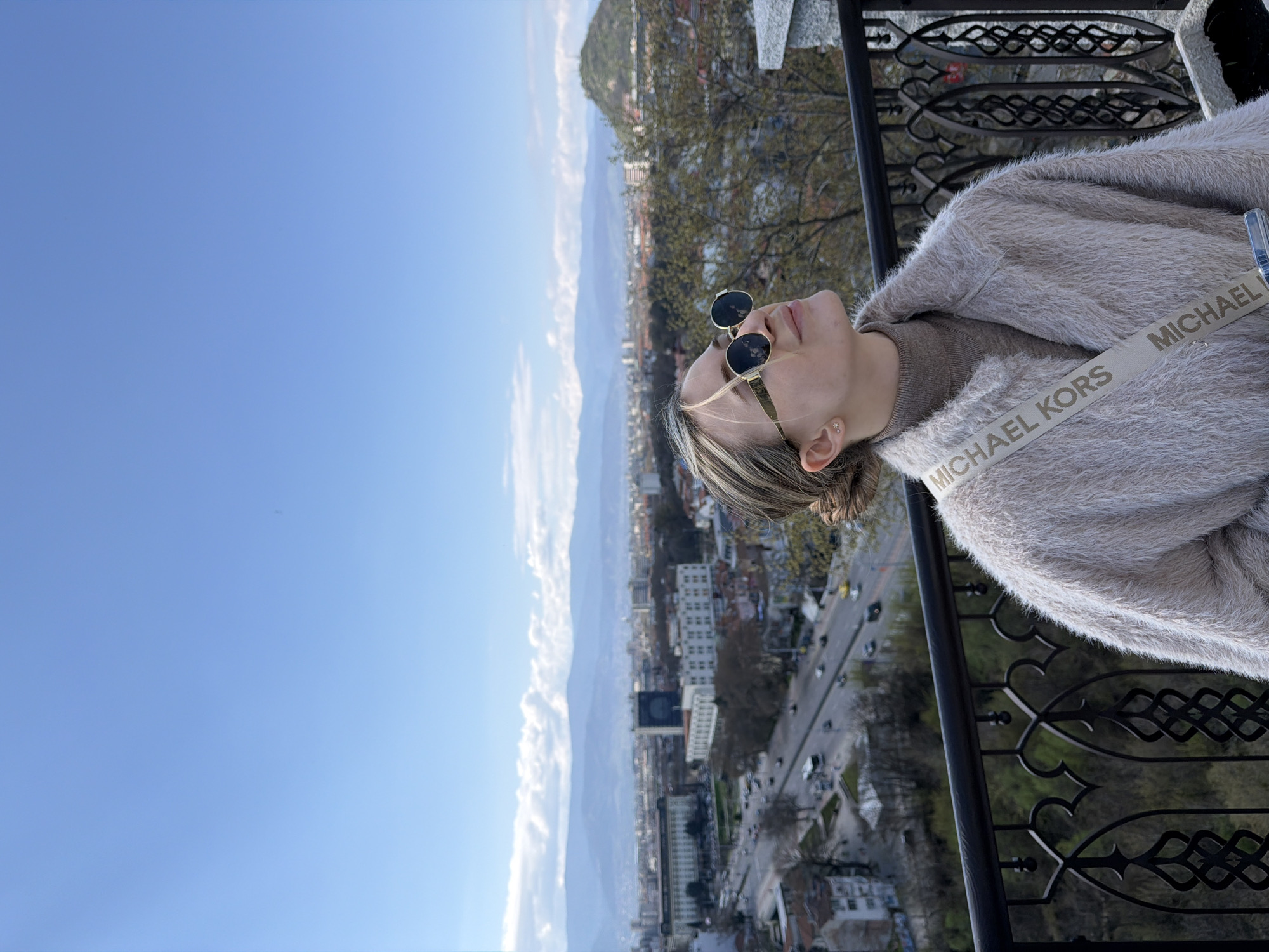 A woman wearing sunglasses and a light-colored, fuzzy coat with a Michael Kors branded strap, is standing on a balcony overlooking a cityscape in Plovdiv, Bulgaria. She is facing towards the left of the image with her head tilted back. The cityscape includes buildings, streets with traffic, trees, and mountains in the background. The sky is blue. A black metal railing is visible in the foreground.