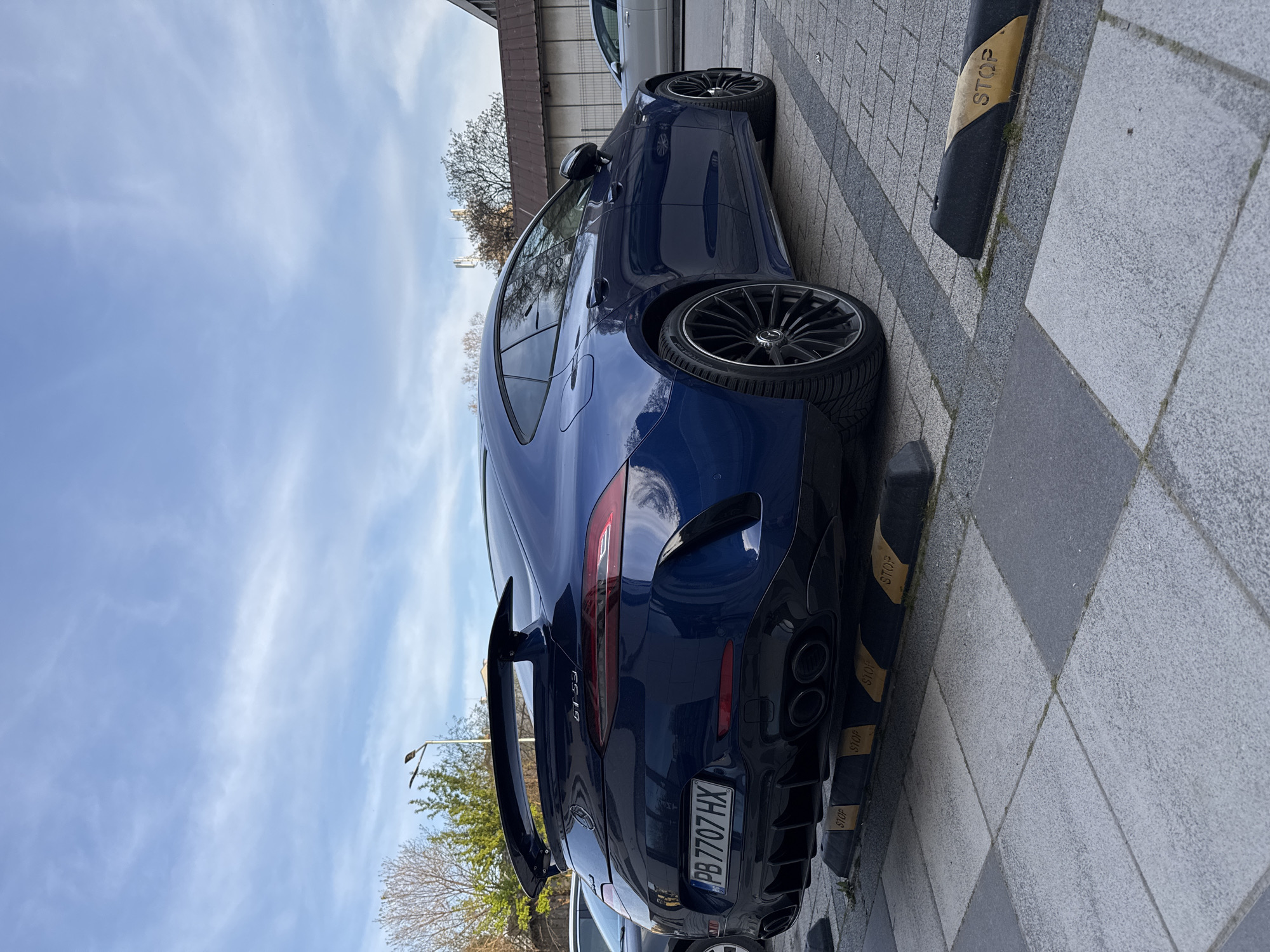 A blue Mercedes-AMG GT 63 S 4-door Coupé is parked on a gray tiled sidewalk. The car has a license plate that reads "PB 7707 HX" and the text "GT 63" is visible on the trunk. The car is positioned next to black and yellow parking stops with "STOP" written on them. In the background, a blue sky with light clouds is visible, along with buildings and trees. Given the license plate and the general environment, the car is likely located in Plovdiv, Bulgaria.