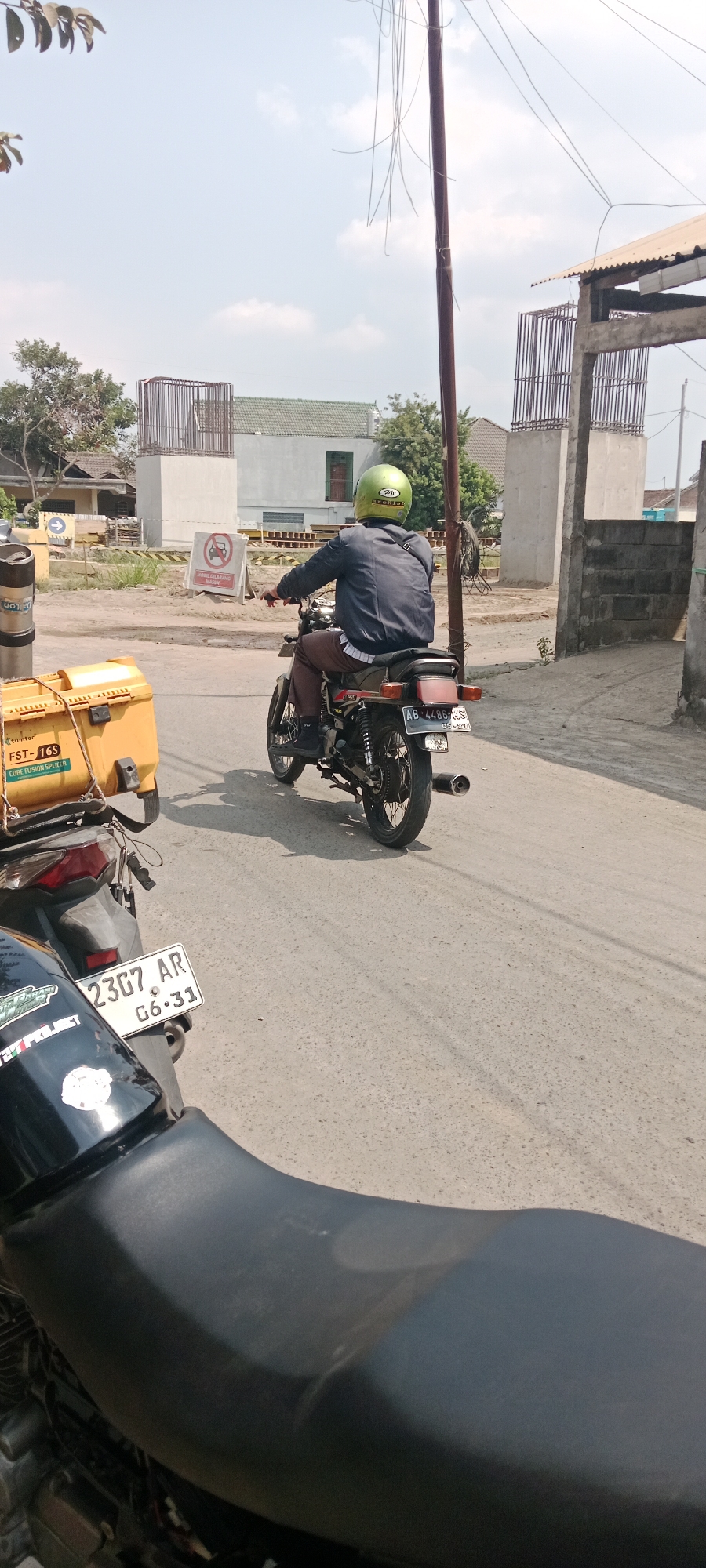 A person wearing a dark jacket and a light green helmet rides a black motorcycle with a silver frame and black seats down a paved road. The motorcycle has a license plate that reads "AB 4486 RS". In the background, there is a building under construction with concrete pillars and scaffolding, as well as other buildings and trees. There is also a yellow container on the left side of the image. The scene appears to be in an outdoor, urban or semi-urban environment, possibly in Purwomartani, Indonesia, indicated by the license plate format. The sky is visible and appears to be bright and clear.