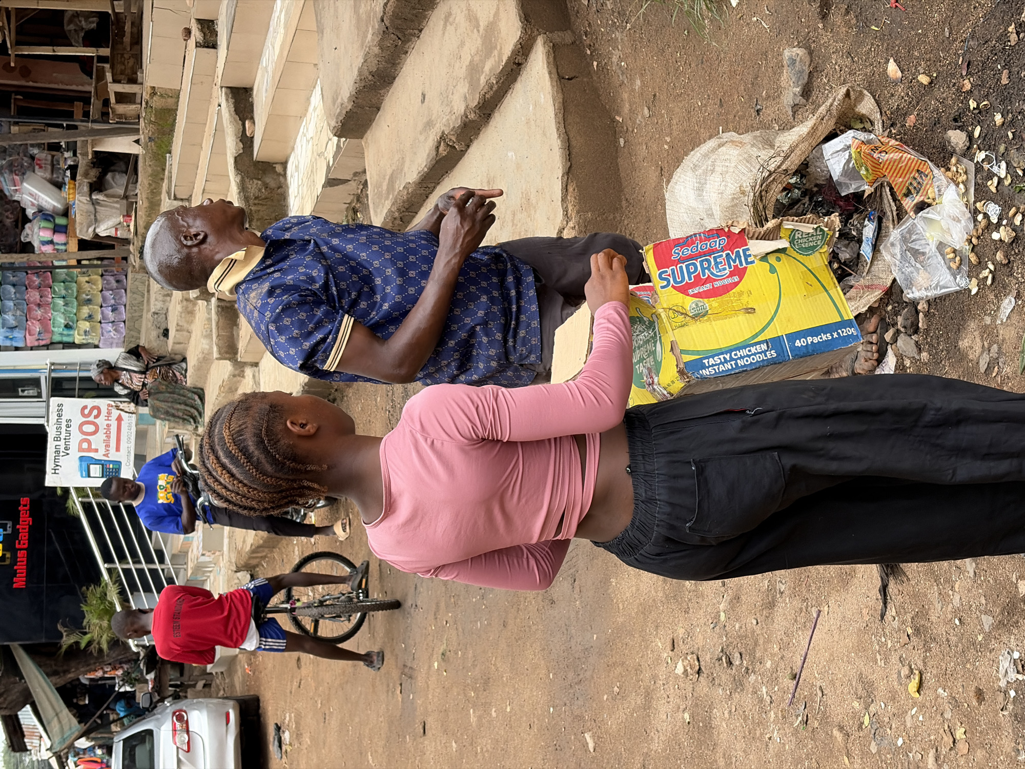 The image appears to be taken outdoors in a bustling, informal setting, possibly in Jos, Nigeria. An older man, with dark skin and a bald head, wearing a patterned blue shirt with gold accents, sits on what seems to be a concrete step, possibly waiting. Next to him, a younger woman with braided hair, wearing a pink long-sleeved crop top and black pants, is interacting with items next to a cardboard box labeled "Sedaap Supreme Instant Noodles - Tasty Chicken."
Beside the woman is a bag full of trash and instant noodle wrappers. In the background, a boy wearing a red shirt rides a bicycle, and another person wearing a blue shirt is near a sign for "Hyman Business Ventures POS" and "Mulus Gadgets." A white car is parked nearby. The ground appears to be dirt or gravel. The atmosphere seems to be a mix of commerce, everyday life, and urban informality.