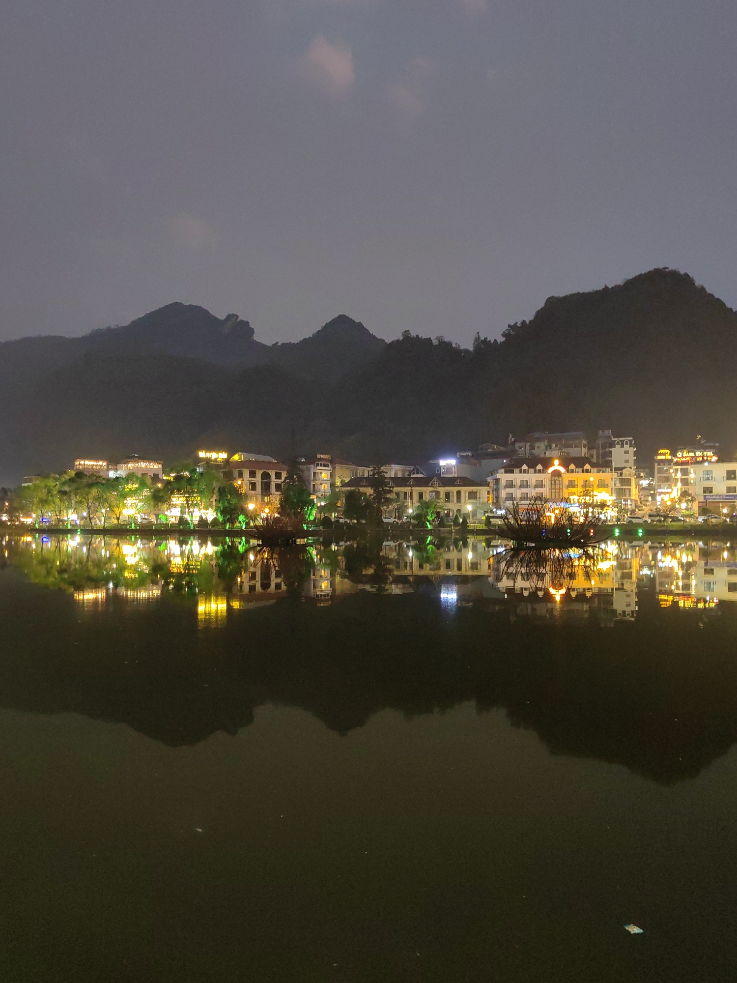 The image depicts Sa Pa Lake in Sa Pa, Vietnam, at night. The foreground consists of the dark, calm surface of the lake, which reflects the illuminated structures and lights from the opposite shore. Along the midground shoreline, a row of multi-story buildings, likely hotels and commercial establishments, display a variety of illuminated windows and facades. Bright yellow and white lights emanate from these buildings, with some signs partially legible, including "SAPA HOTEL" and "LUXURY TRAVEL." Trees line the lake's edge, also illuminated with green and yellow lights. A prominent, dark, decorative structure resembling a stylized lotus flower, adorned with various colored lights, floats on the lake near the shore. The background features dark, silhouetted mountains rising against a hazy, dark gray night sky, where faint hints of reddish-orange clouds are visible at the top. No individuals or specific actions are discernible. The scene is characterized by artificial illumination reflected on the water, creating a symmetrical visual effect.