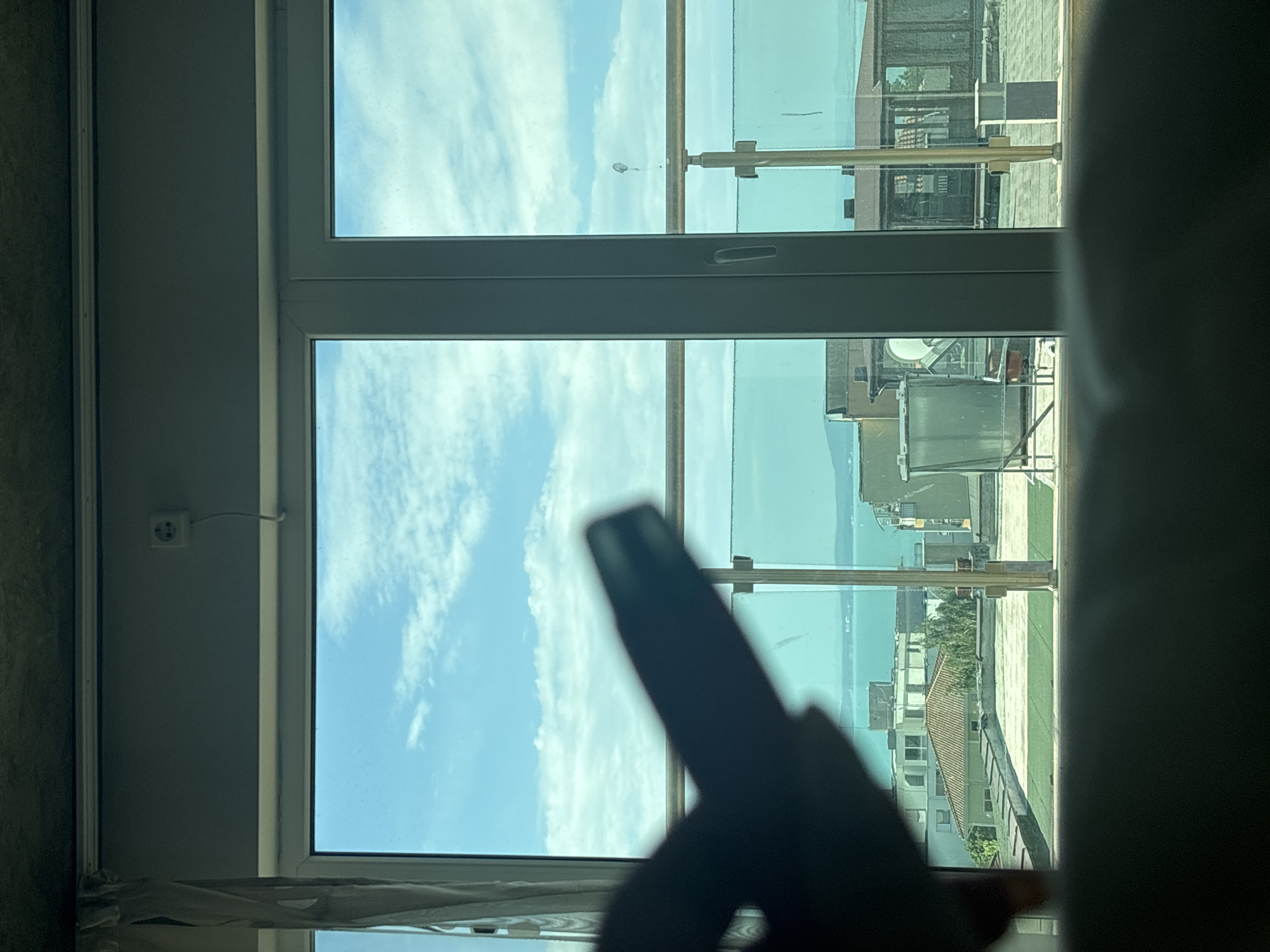 The image shows a view from inside a building, looking out through a window. The window is divided into three panes, with a horizontal bar and vertical supports. Through the window, a landscape is visible, featuring a body of water, possibly a lake or sea, and the skyline of what appears to be a coastal city. The sky is blue with scattered clouds.
The scene outside the window includes rooftops, buildings, and structures that suggest an urban environment. Some rooftops have what appear to be metallic or reflective surfaces. The architectural style of the buildings is modern.
In the foreground, within the building, part of a white wall or curtain is visible. A silhouette of a hand holding a vaping device is visible in front of the window. The device is held up against the skyline, partially obscuring the view.
Based on the information in the prompt, the location of the scene outside the window is Burgas, Bulgaria.