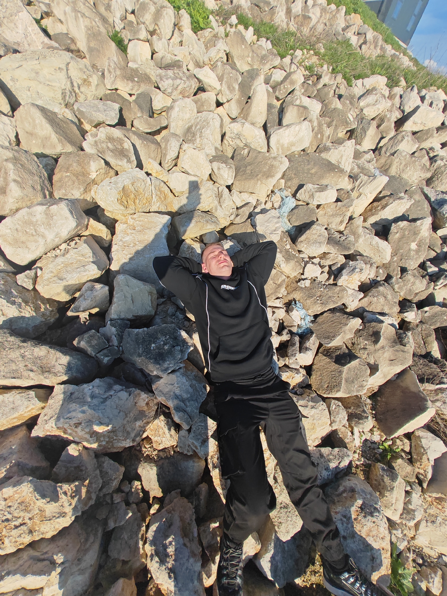A man is lying on a pile of large, light-colored rocks. He is wearing a black hoodie with white drawstrings and black pants and shoes. His hands are behind his head. The background is a bright blue sky. There is green grass growing between the rocks, and a building is visible in the upper right corner. The scene appears to be outdoors in a sunny location. The location is possibly in the city of Shumen, Bulgaria.