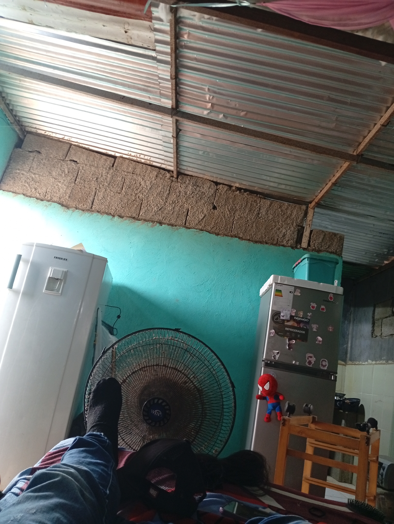 This image captures an interior scene within a residence in Altagracia, Venezuela, taken from a low perspective, consistent with a person lying down. The lower body of an individual, clad in blue jeans and dark socks, rests on a striped textile in the bottom-left foreground. A black baseball cap bearing "MARVEL" branding and a dark-colored hairbrush are visible near the individual's feet.
The room features an exposed roof structure composed of corrugated metal sheets supported by rough wooden beams and minor structural elements. A small section of pink fabric is draped over a beam in the upper right. The walls exhibit a two-tiered construction: the upper portion consists of exposed, unrendered concrete blocks with visible mortar, while the lower sections are painted a vibrant turquoise.
Household appliances are prominent. On the left, a white "FRIGILUX" brand single-door refrigerator is positioned. A large, black "WIND'S" brand stand fan with a metal grille is centrally located in the foreground, facing the viewer. On the right, a silver-grey "DAEWOO" brand two-door refrigerator is visible, displaying an energy efficiency label, numerous magnetic decorations, and a red Spider-Man plush toy attached to its lower door. A light-blue plastic storage bin rests on top of this refrigerator. A simple wooden chair or stool with visible slats is situated to the right of the silver-grey refrigerator. A background wall of unpainted concrete blocks is partially visible behind the right refrigerator and chair. The room is illuminated by ambient natural light.