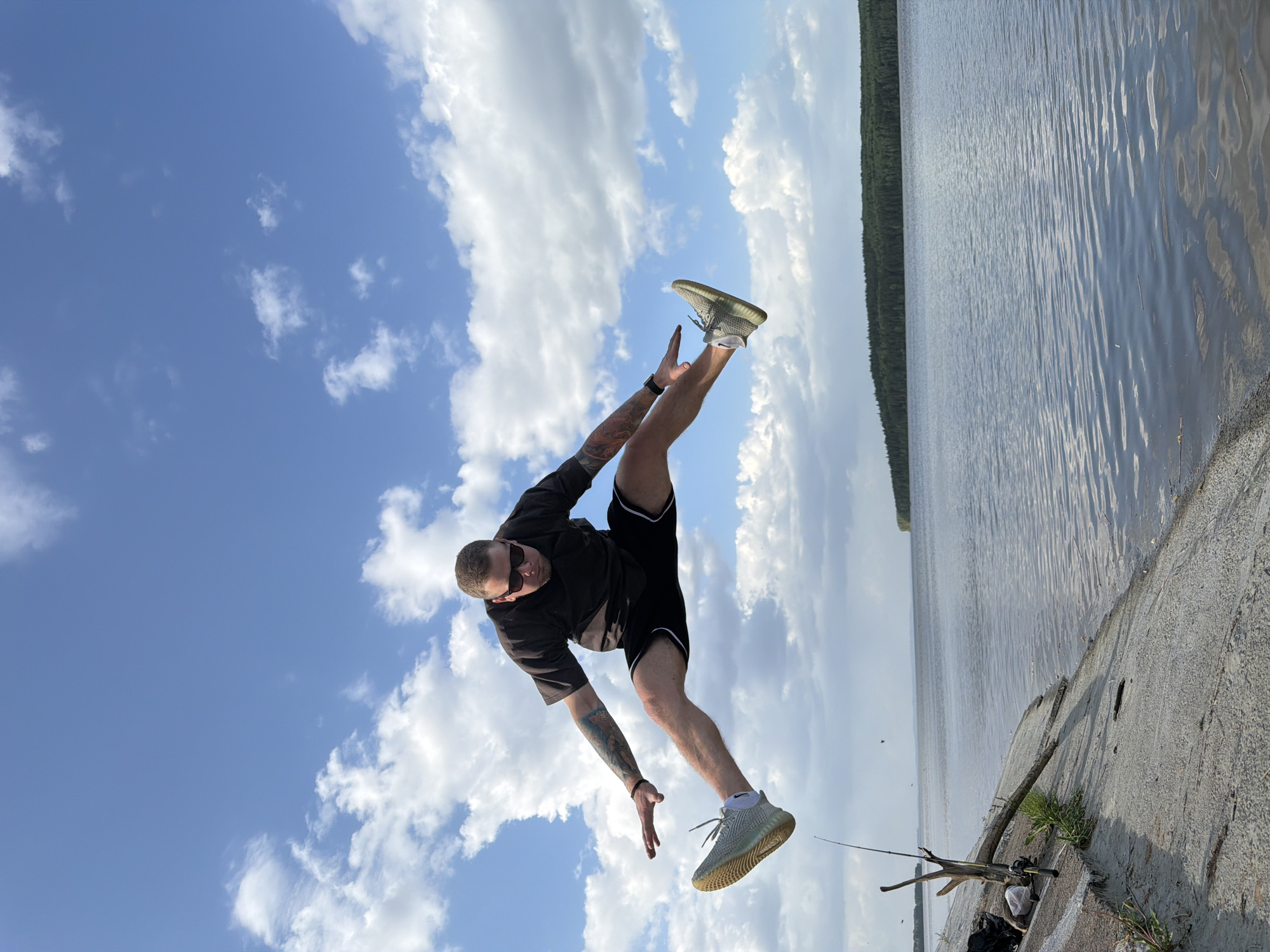 A man is captured in a dynamic pose against a backdrop of a body of water, likely a river or large lake, and a sky filled with fluffy white clouds. He is wearing black shorts and a black short-sleeved shirt, sunglasses, and grey athletic shoes. His right arm is extended upward, while his right leg is raised high in the air. The position of his body suggests motion or a playful moment.
The scene is situated near a concrete embankment, where fishing poles are visible, indicating that fishing is a possible activity in the area. The location is identified as Chaikovsky, Russia, due to the request in the prompt. The environment features a blend of natural elements, such as the water and sky, with the man-made structures of the concrete embankment and fishing equipment.