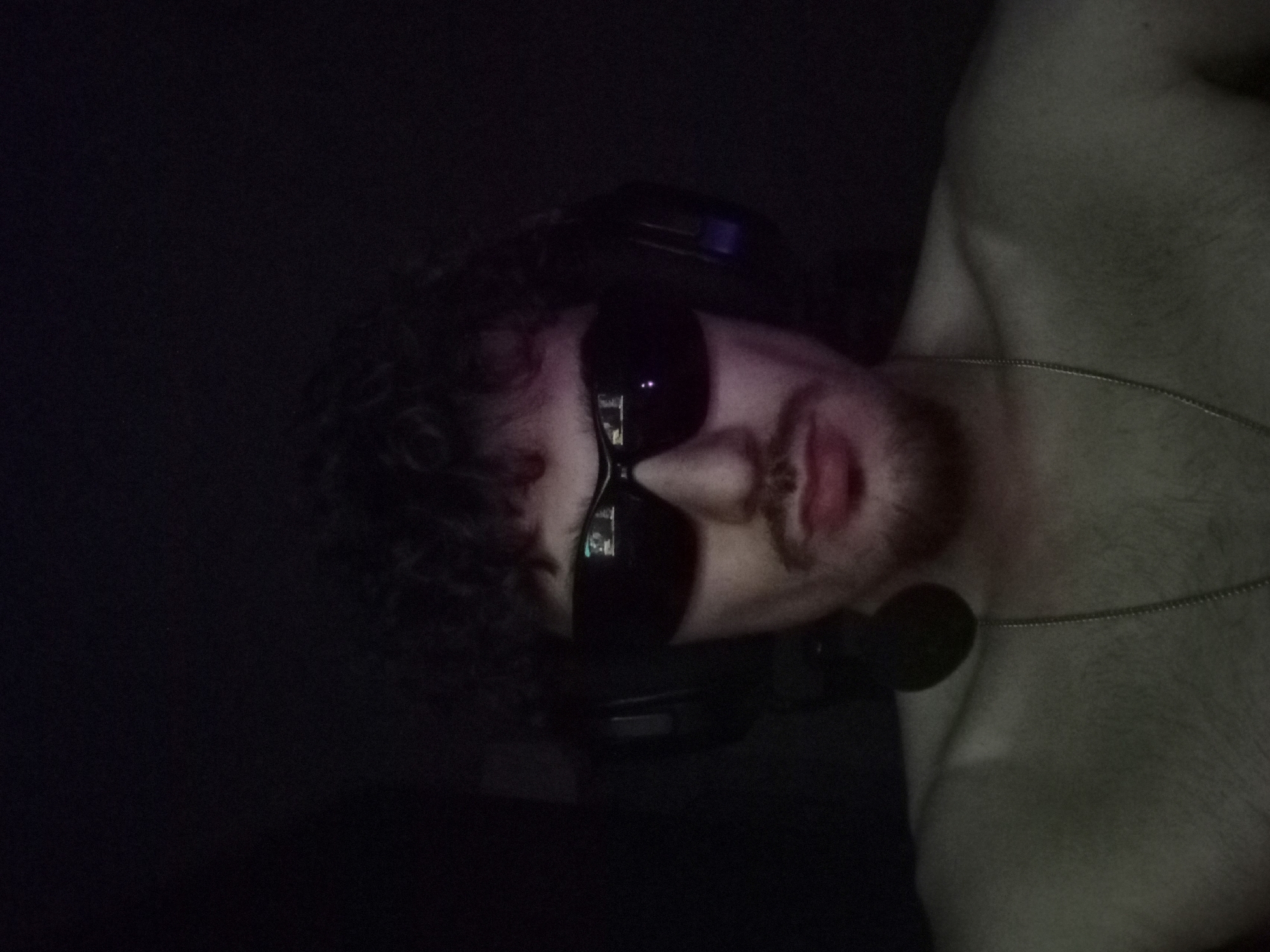 The image depicts a close-up, frontal view of a young man with curly dark hair and a mustache, wearing black sunglasses and headphones. He is shirtless, revealing his shoulders and chest, with a silver chain necklace visible. The lighting is dim, creating a dark environment with subtle highlights on his skin. The background is almost entirely black, with some faint texture suggesting an indoor setting. There is no discernible information about the location in Parvomay, Bulgaria, as the scene is highly focused on the subject and lacks any contextual environmental details specific to that city.