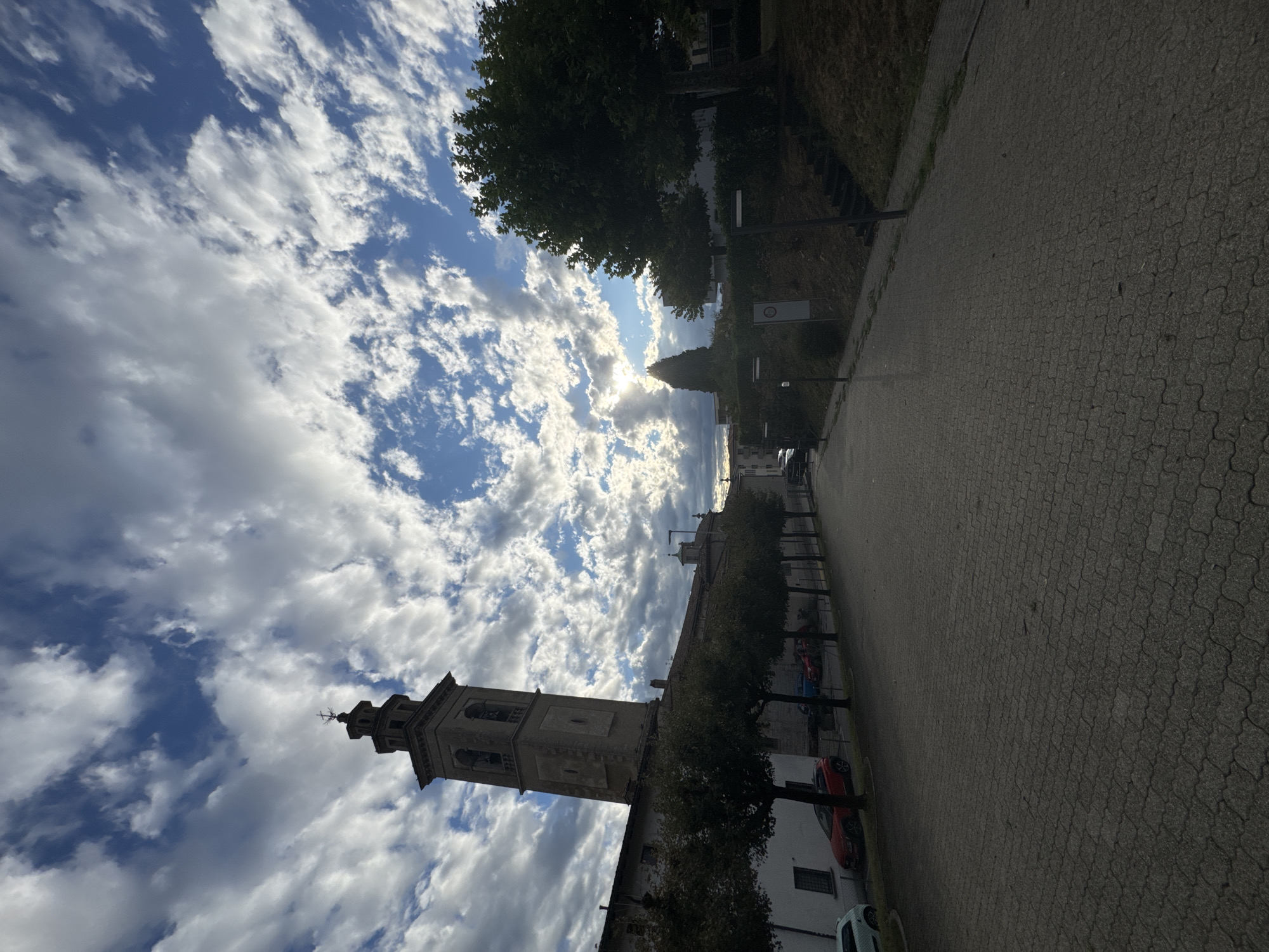The image depicts a street scene in Balerna, Switzerland, dominated by a tall, slender bell tower of a church or similar structure. The tower is made of stone and features arched openings and a cross at its apex. To the left of the tower, a long building with a white facade stretches along the street, partially obscured by a row of evenly spaced trees with dark green foliage. Parked along the side of the street in front of this building are several cars, including a red SUV. The street itself is paved with interlocking grey pavers, and on the right side of the street, a grassy embankment rises, topped with trees and a dark green bush. Several modern-style lampposts are visible along the street and on the embankment. The sky above is filled with dramatic, white and grey cumulus clouds against a bright blue sky, with the sun partially obscured by the clouds, creating a bright glare in the center of the frame. The overall impression is of a quiet town square or street on a day with dynamic cloud formations.