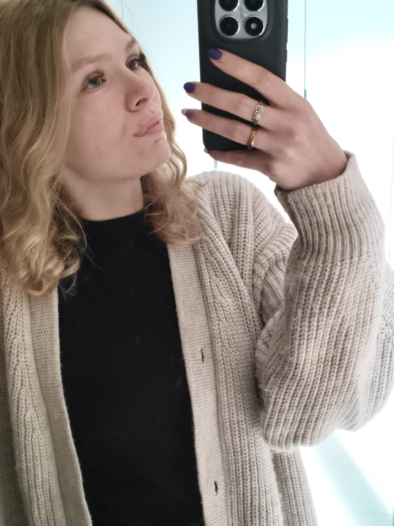 A woman is taking a selfie in a room with a blue-toned wall. She is wearing a black top and a light beige, button-down cardigan. Her blonde hair is styled in loose waves. She is holding a black smartphone, her fingers visible, displaying purple nail polish and two rings – one silver with a decorative pattern and another a simple gold band. The location is not discernible.