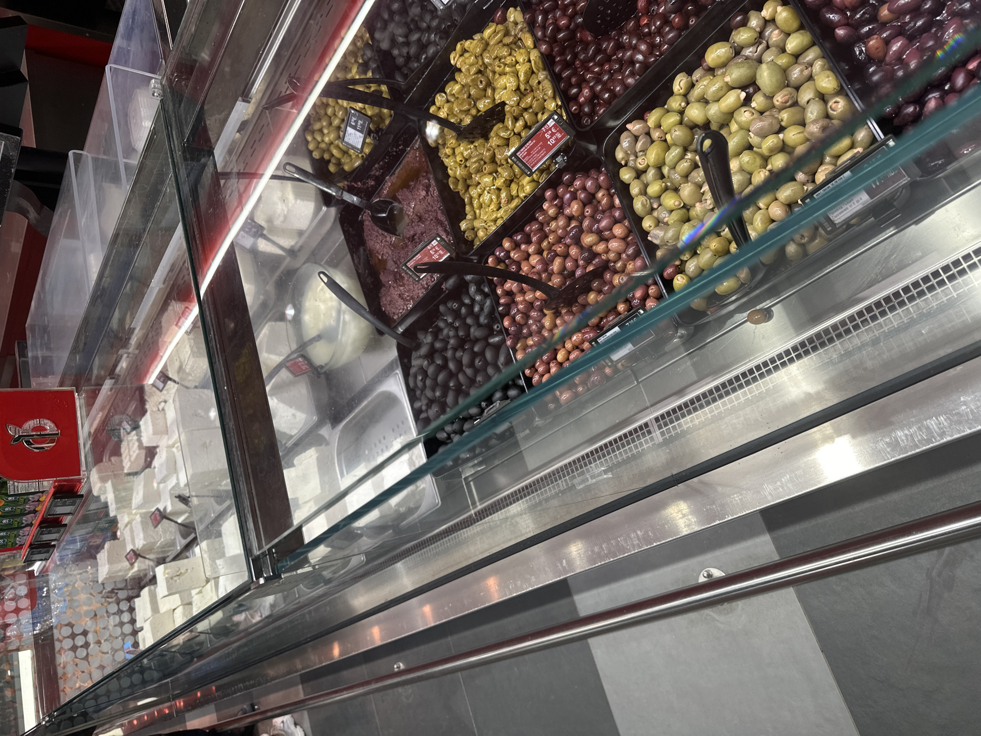 This image provides an overhead view of a commercial food display case, likely in a deli or supermarket in Sofia, Bulgaria. The display is separated by clear glass partitions and features a variety of olives and fresh cheese.
On the right side of the case, numerous large bins are filled with different types of olives. These include vibrant green olives, dark black olives, and reddish-brown olives, suggesting various curing methods. One bin also appears to contain a dark, crushed olive preparation, possibly tapenade. Black serving scoops are placed within most of the olive containers, indicating a self-serve or assisted-serve setup. Several price tags are visible on the bins, displaying costs in Euros, such as "5.59 € / 100g", "6.29 € / 100g", and "1.79 € / 100g".
On the left side of the display, multiple rectangular blocks of white cheese, consistent with feta or a similar fresh cheese, are neatly stacked in clear compartments. A red container with a white, stylized fish or symbol logo is partially visible towards the far left. The display case itself has a clean, polished stainless steel base and a metal railing along the front. The environment appears well-lit, and the food items are presented in an organized manner. No people are visible in the image.