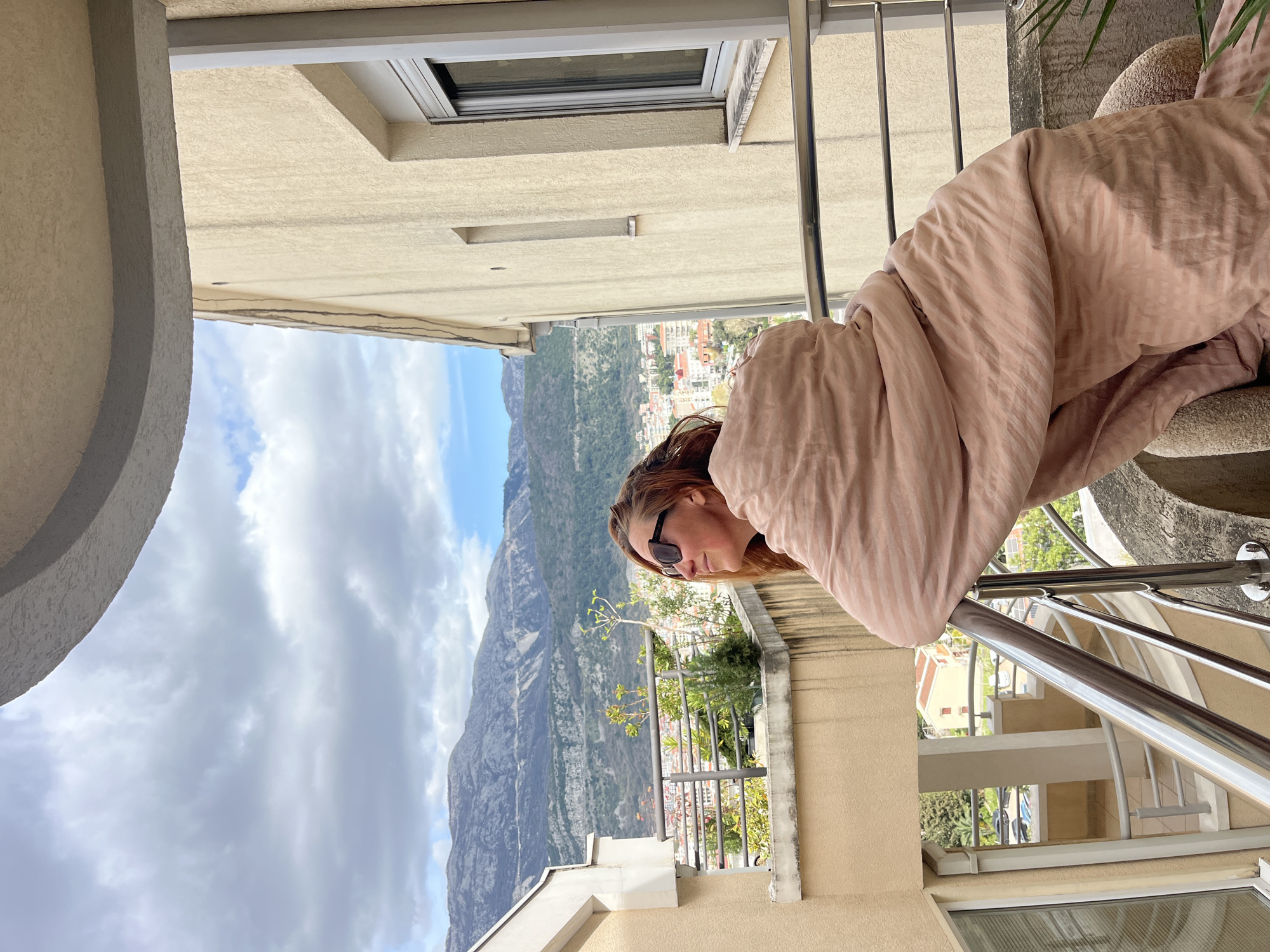 A woman wearing sunglasses and wrapped in a striped, peach-colored blanket looks out from a balcony. The balcony has a stainless steel railing. Below, there is a view of the mountainous landscape and buildings of Budva, Montenegro. The sky is partly cloudy.
