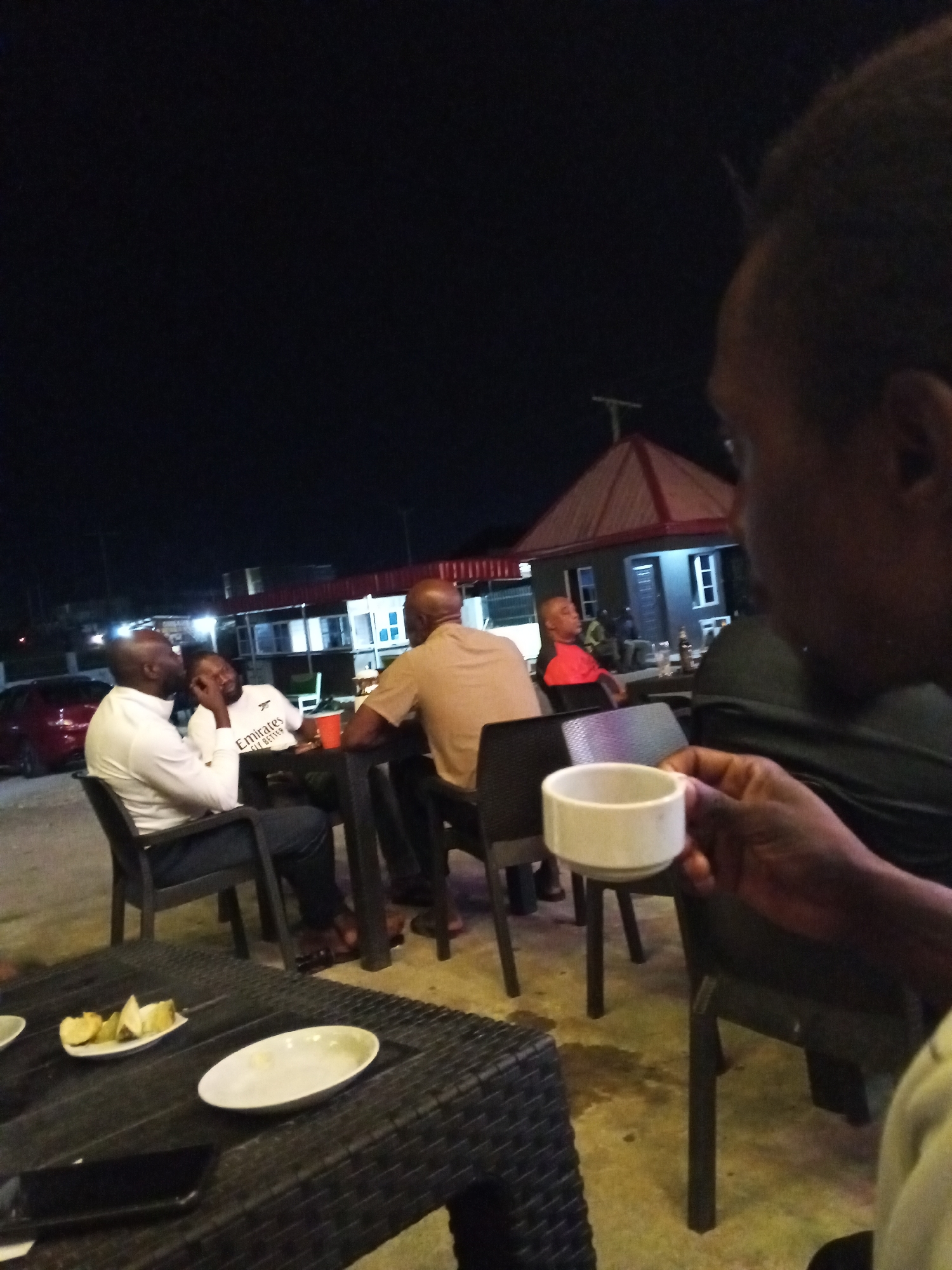 At night in Lokoja, Nigeria, several people are gathered outdoors. A group of men sit at tables and appear to be engaged in conversation or socializing. One person is holding a white cup, and on the table in the foreground are plates with food remnants and a mobile phone. The background reveals a building with a red-roofed structure and lit windows. A car is parked nearby. The overall scene suggests a casual, social gathering in an outdoor setting.