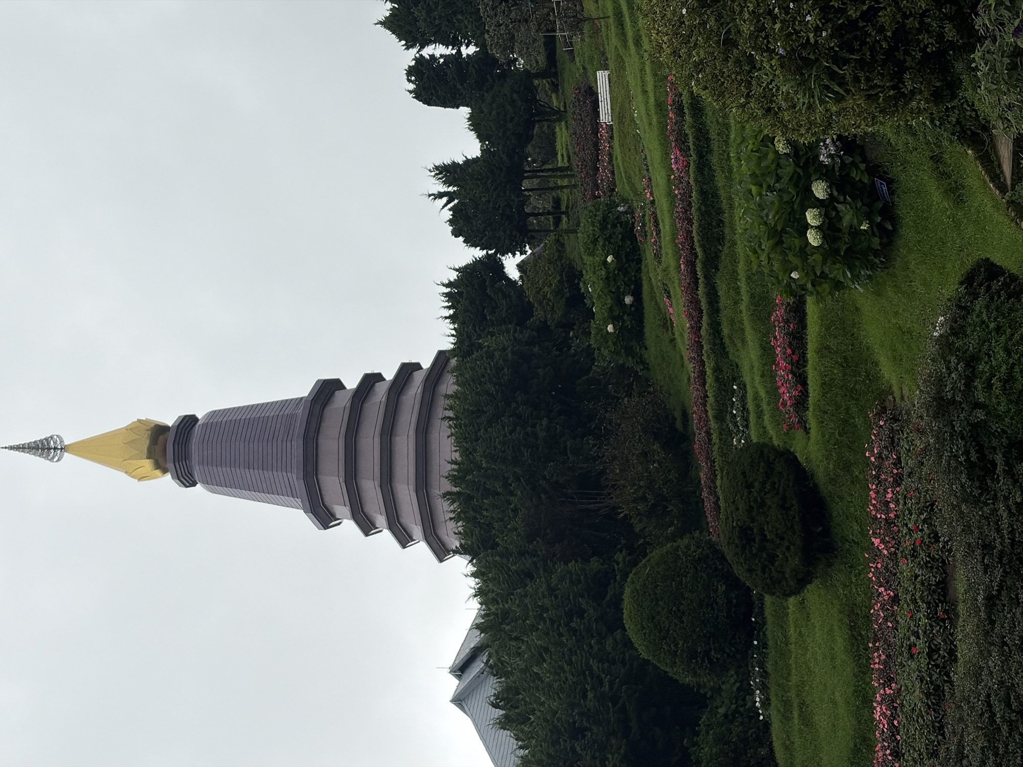The image depicts a tiered, stupa-like structure with a golden, pointed finial, set against an overcast sky. The stupa is flanked by a pitched-roof building on the left and surrounded by lush greenery. In the foreground, a meticulously landscaped garden features rolling hills covered in grass, with terraced beds of flowers in various shades of pink and red. Several rounded, dark green bushes are strategically placed on the grassy slopes. In the middle ground, a white bench is visible on one of the terraces. The background is filled with dense evergreen trees. The overall scene suggests a temple or a well-maintained botanical garden. This structure is likely the Phra Mahathat Naphamethanidon and Phra Mahathat Naphamongkol at Doi Inthanon National Park in Chom Thong District, Chiang Mai Province, Thailand, which is located in the subdistrict of Tambon Ban Luang. There are no people or animals visible in the image.