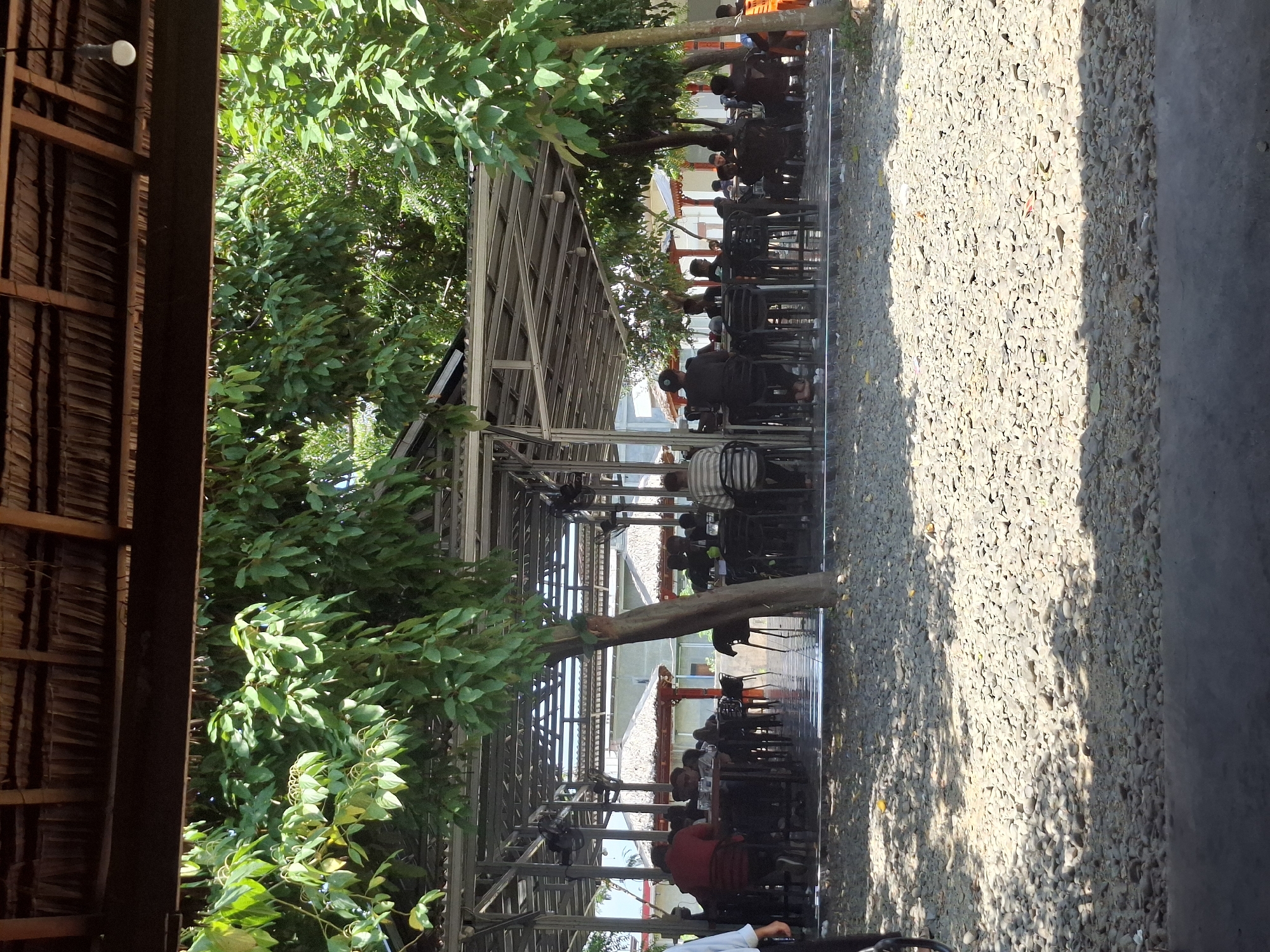 The image depicts an outdoor dining area of a restaurant, possibly located in Keuniree, Indonesia, given the context. The structure features a thatched roof supported by metal beams, providing shade. Below, numerous tables and chairs are arranged, with several patrons seated and seemingly engaged in dining or conversation. The ground is covered in small, light-colored pebbles. Lush green trees and foliage surround the dining area, contributing to a natural, open-air ambiance. The overall scene suggests a casual and relaxed setting.