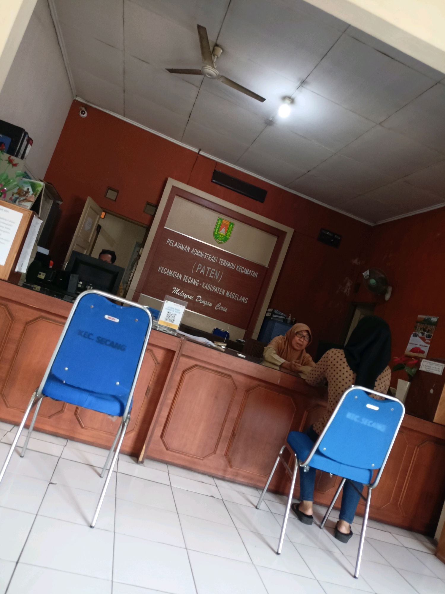 The image depicts the interior of a government office, likely a service center, in Kewaluhan, Indonesia. A wooden counter dominates the foreground, behind which two women are visible. One woman, wearing a hijab and an orange headscarf, is seated at the counter, appearing to interact with the other woman. The second woman, also wearing a hijab and a polka-dotted top, stands behind the counter.
In front of the counter, two blue plastic chairs with metal frames are positioned. One chair is closer to the viewer and has "KEC. SECANG" printed on its backrest. The walls of the office are painted a reddish-brown color, and a large sign with text in Indonesian, likely indicating the office's name and services, is mounted on the wall. A ceiling fan and a light fixture are visible on the white ceiling. The floor is tiled in a light color. The overall impression is of a functional, if somewhat dated, administrative environment.