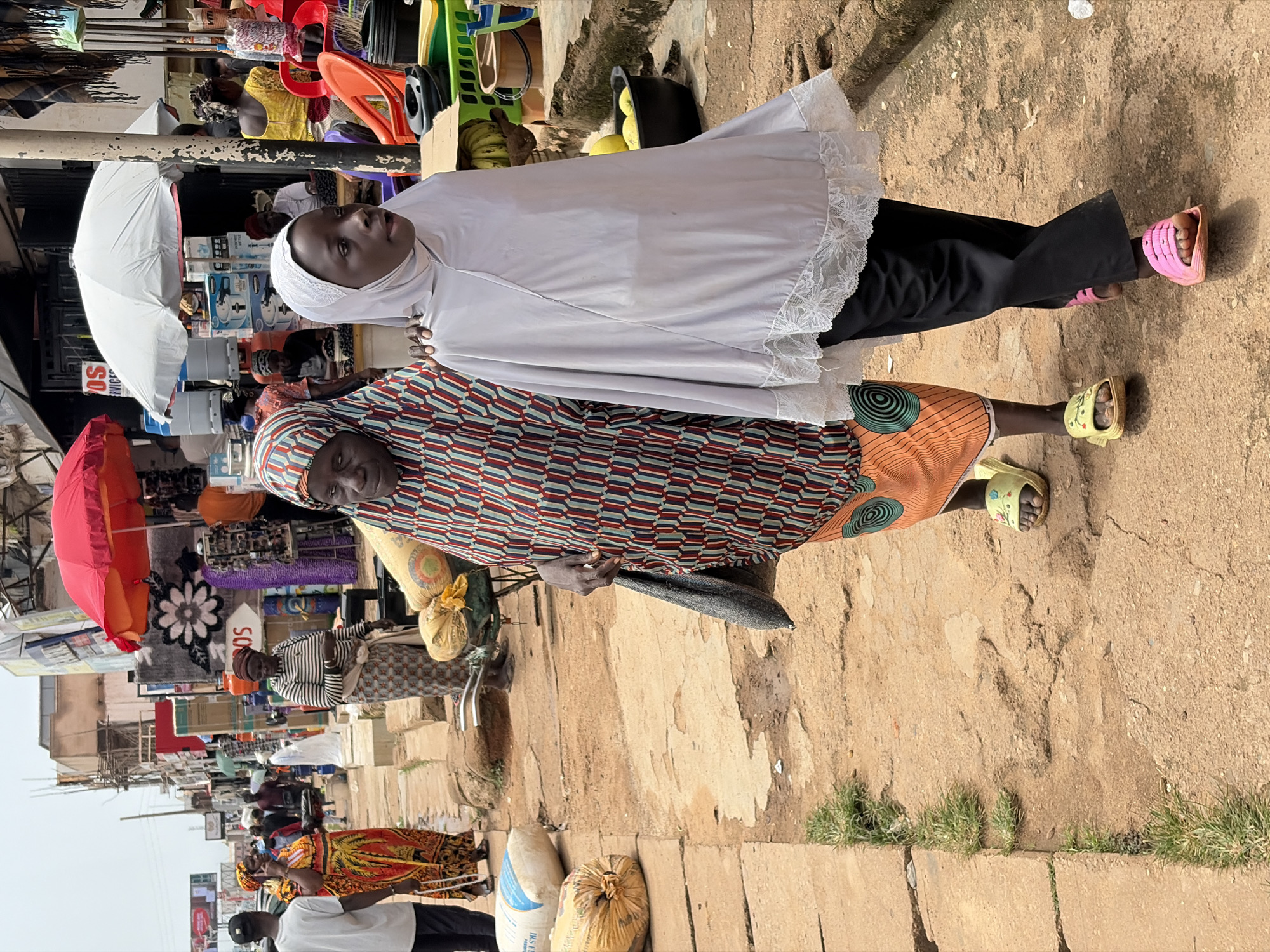 Two women, one older and one younger, are walking side-by-side on a dirt path in an outdoor market. The older woman wears a vibrantly patterned headscarf and a dress with an orange and red striped pattern. She is carrying a dark fabric bag over her shoulder. The younger woman is wearing a light-colored hijab and black pants. She has on pink slides, and the older woman has on yellow slides. In the background, there are market stalls with various goods, including textiles, umbrellas, and food items. Several other people are visible in the background, some shopping and others working. The environment appears to be a bustling street market in Jos, Nigeria, characterized by its unpaved ground and the array of vendors and shoppers.