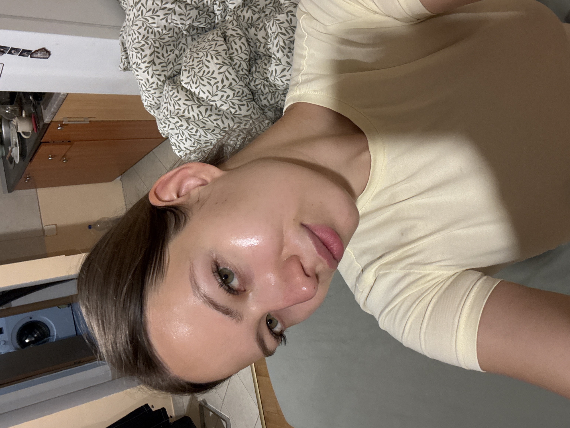 A woman with fair skin and olive green eyes poses in an indoor location. She wears a light yellow t-shirt and has dark brown hair. Behind her, there is a bed with a leaf-patterned comforter. To the left, kitchen cabinets and a washing machine are visible. The location is in a home. There is no specific information available to indicate the location is in the city of Bečići, Montenegro.
