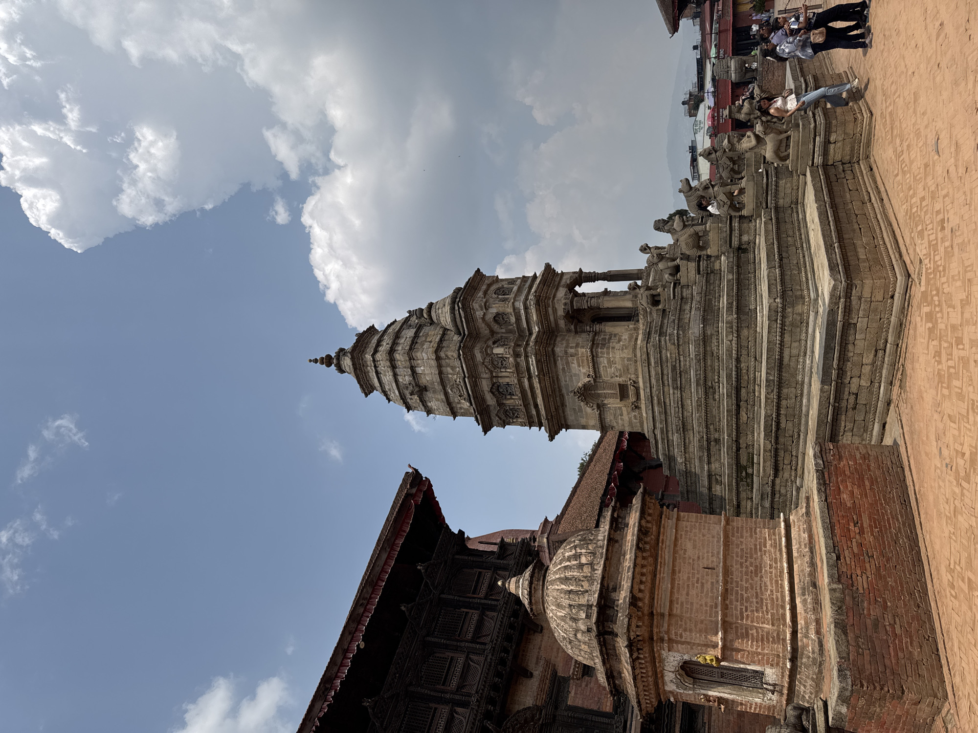 The image shows the Nyatapola Temple in Bhaktapur, Nepal. The five-story pagoda-style temple rises prominently against a blue sky with scattered clouds. Below and slightly offset is another building with intricate dark wood carving details. The temple is constructed from brick and wood with detailed carvings, and stone steps lead up to the temple. On the steps, there are tourists and locals. The surrounding area is paved with brick.