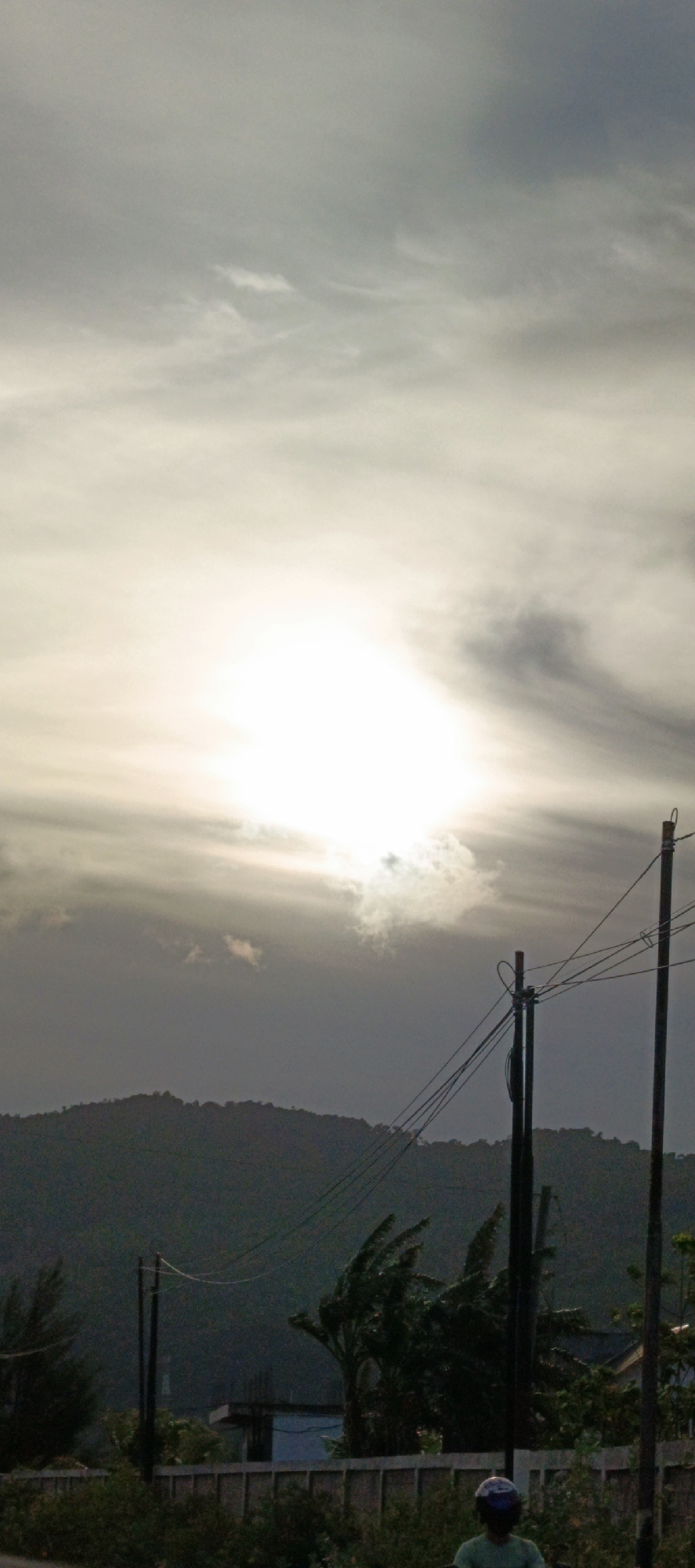 The image captures a hazy, overcast sky with the sun faintly visible behind the clouds, suggesting either early morning or late afternoon light. In the foreground, a silhouette of a person on a motorcycle is partially visible at the bottom right, wearing a blue helmet. Utility poles with wires are prominent throughout the mid-ground, dissecting the landscape. Behind the poles, a dark, undulating line of hills or mountains forms the backdrop, covered in dense foliage. The lower portion of the image shows a fence line with various types of vegetation and what appears to be a partially constructed building. The scene is set in a rural or suburban area, with the environment suggesting a humid, possibly tropical climate, consistent with Lamguron, Indonesia.