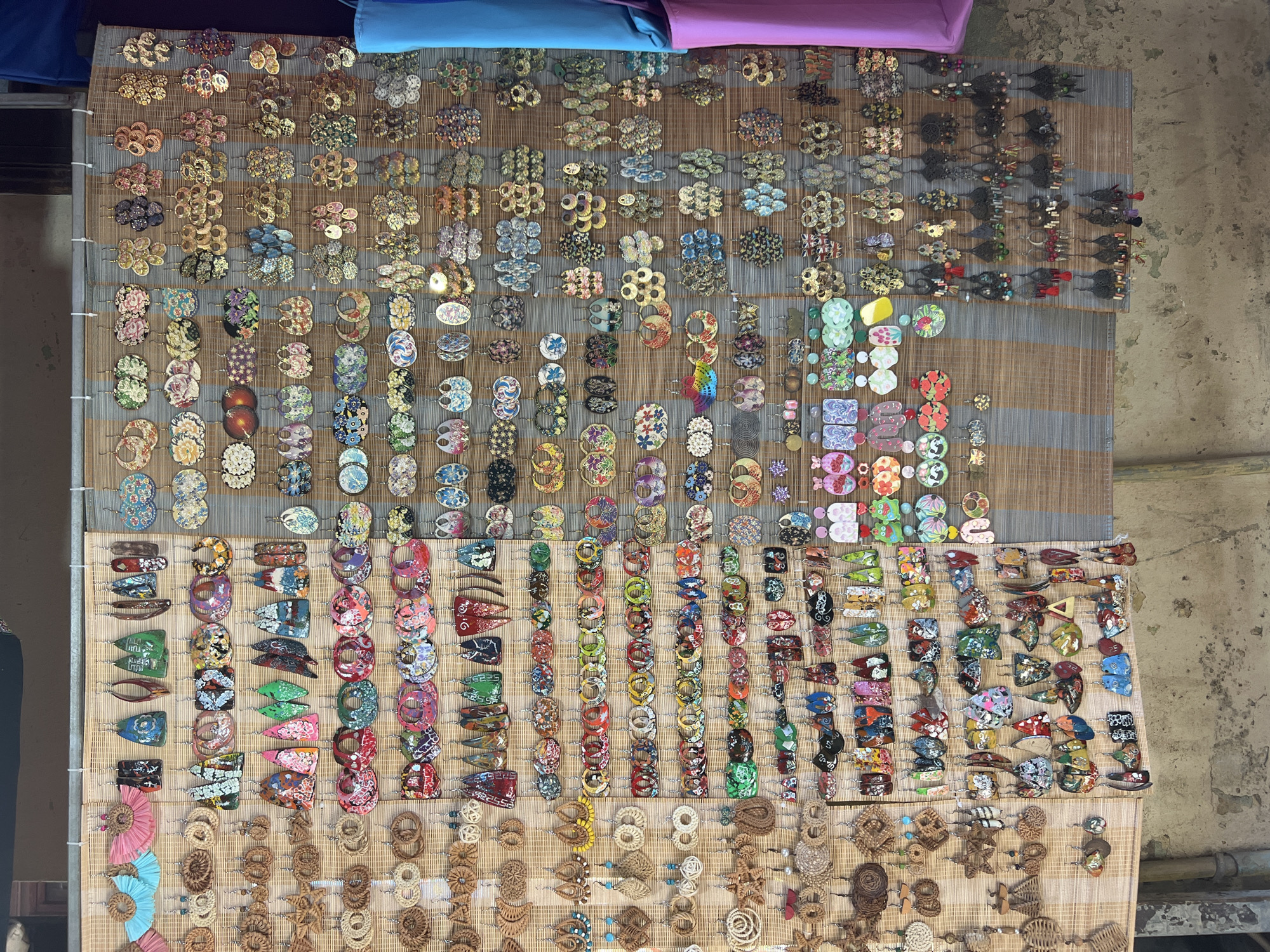 The image shows a stall selling a large assortment of handmade earrings at a market in Hội An Ward, Vietnam. The earrings are displayed on a woven bamboo mat. The earrings are diverse in design, material, and color, with many featuring intricate patterns, natural elements like shells and wood, and vibrant colors. They are hung in neat rows, creating a visually appealing presentation. The background of the image suggests an outdoor market setting, with a rough concrete floor and a hint of a building structure. There are no people visible in the frame, focusing the attention solely on the merchandise. The lighting appears to be natural daylight, highlighting the details of the earrings.