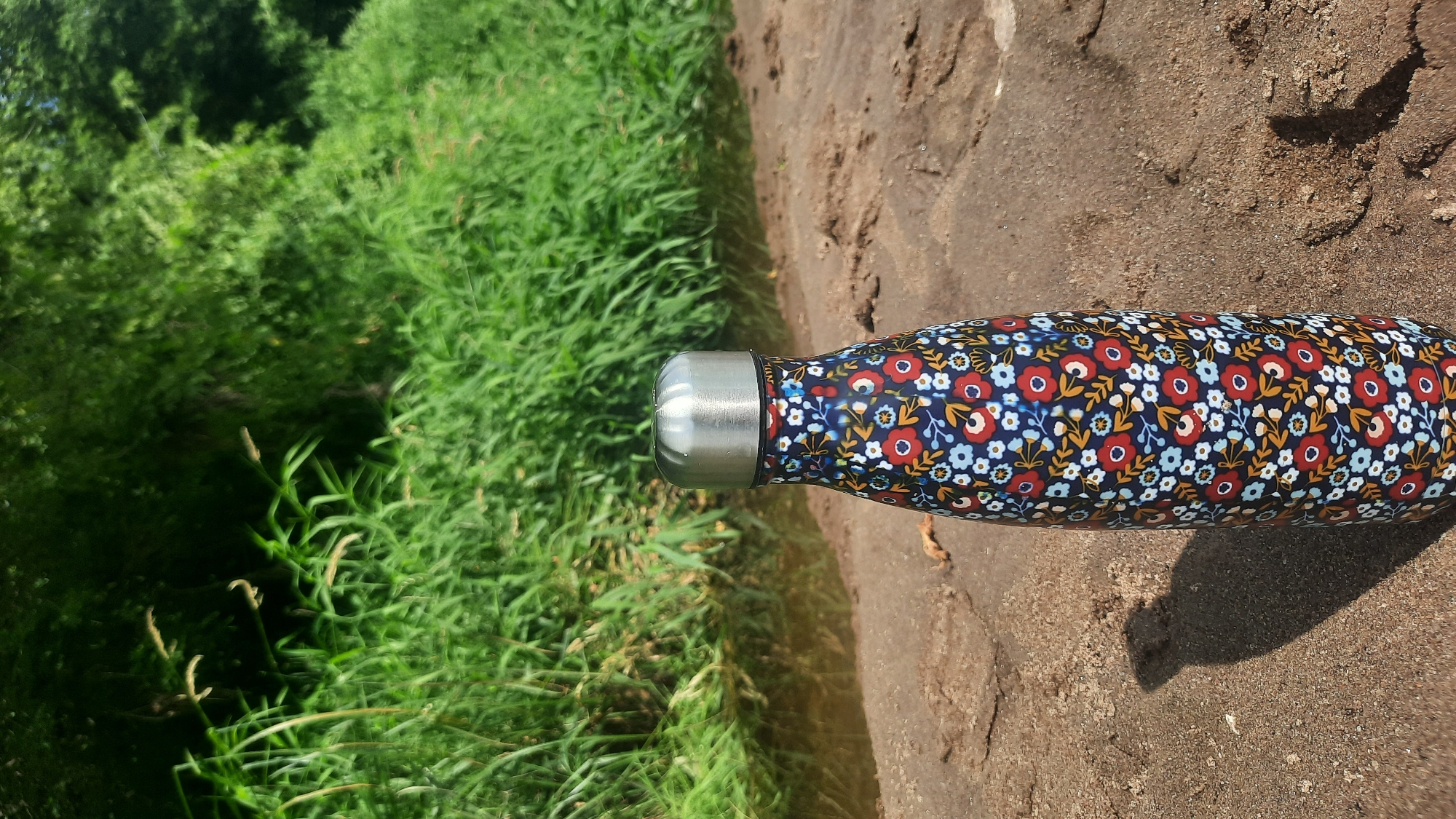 A close-up, low-angle shot focuses on a patterned reusable water bottle resting on sandy ground near a body of water. The bottle, adorned with a blue background and a floral pattern of red, white, and yellow flowers with green leaves, stands upright. Its metallic silver cap is visible at the top. The ground is composed of sand with visible footprints and debris. Behind the bottle, a narrow, calm stream or ditch reflects the sky. Lush green grass and foliage line the far bank of the water. The background is dominated by dense green trees and vegetation, suggesting a natural, possibly wooded environment. The lighting indicates a sunny day, casting a shadow of the bottle to its left. The location is specified as Shklow, Belarus.