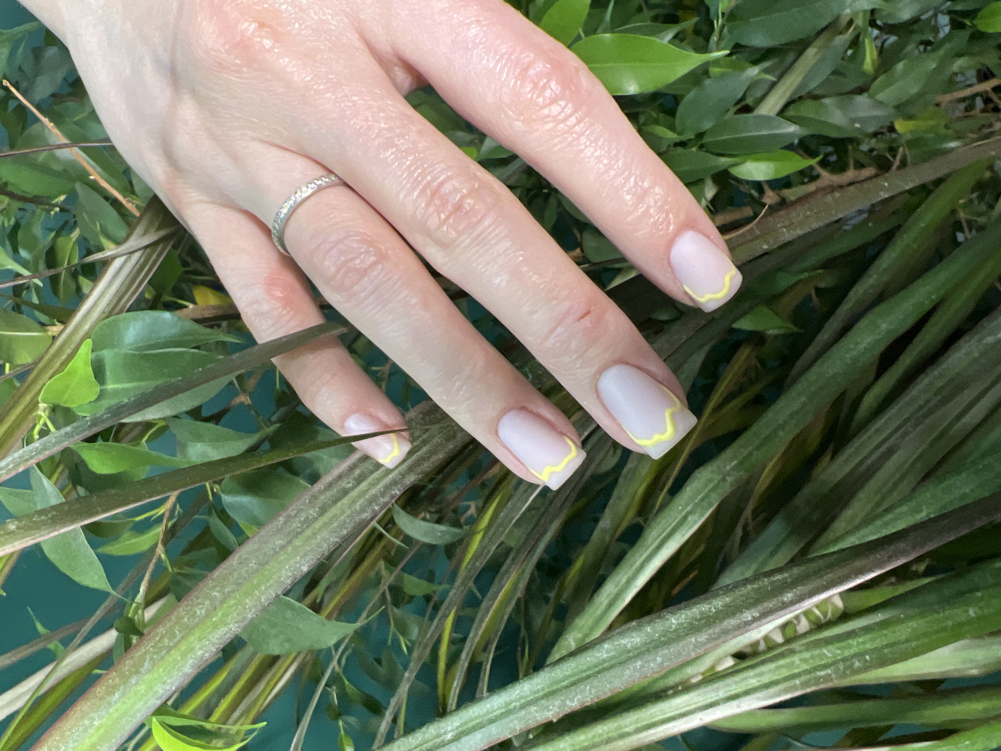 A right hand with fair skin and short fingernails rests on a bed of green leaves and foliage. The fingernails are painted a light, neutral color with a wavy, yellow design along the tips. The index finger has a silver ring with embedded stones. The hand is positioned diagonally across the frame, with the fingers extended. The background consists of various green leaves and stems, some with a variegated pattern of green and lighter yellow or beige. The image appears to be taken indoors, possibly in a setting with decorative greenery.