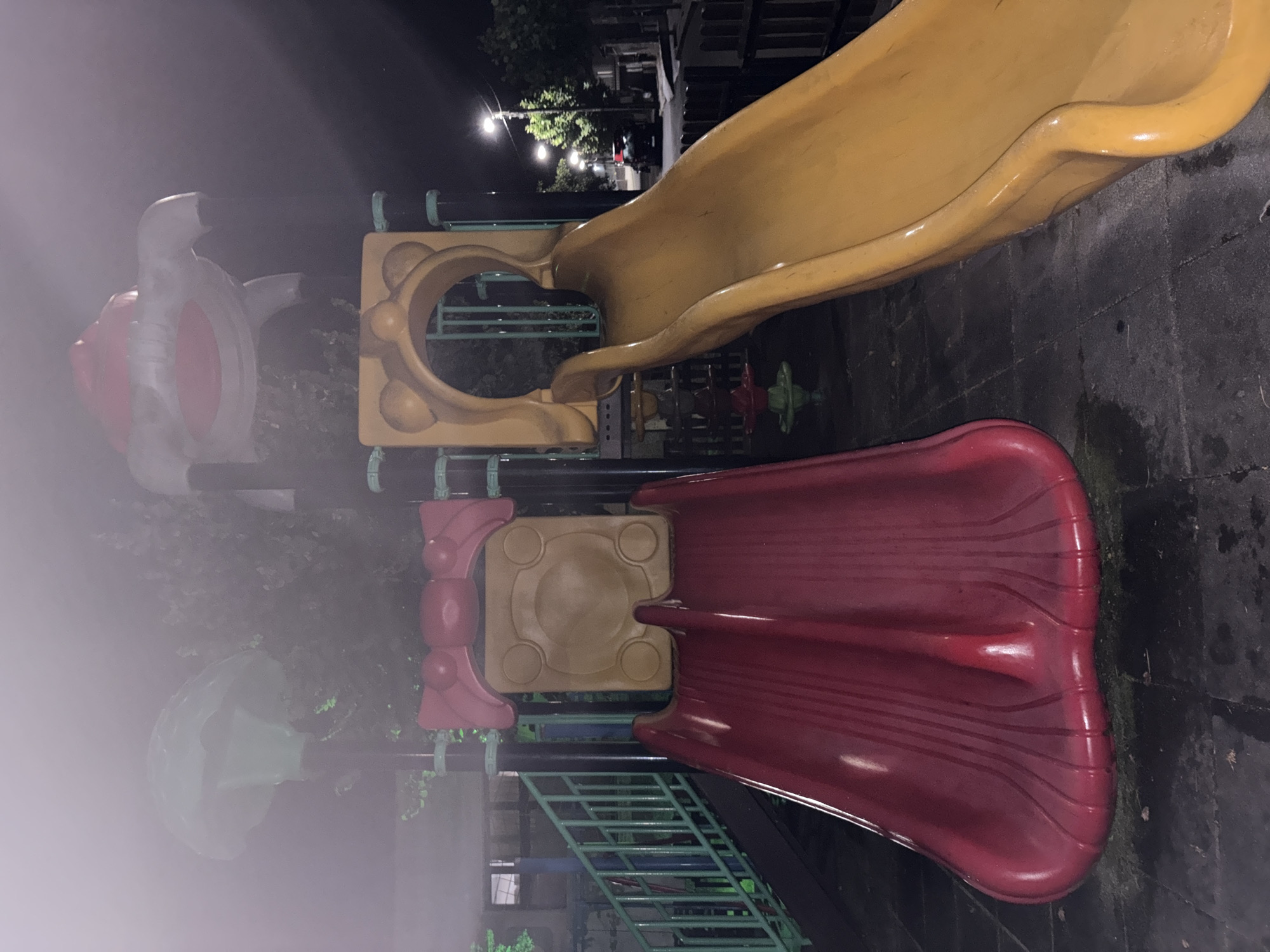 This is a nighttime, medium-shot, eye-level photograph of a playground with two slides in Obzor, Bulgaria. The playground is illuminated by streetlights, and a mist or fog is present, creating a soft, hazy atmosphere. On the left, a red slide descends from a platform adorned with a red bow-like structure and a circular yellow panel. To the right, a yellow slide curves downward from a yellow structure with ear-like protrusions. In the background, a green slide structure and what appears to be a mushroom-shaped object are visible, also partially obscured by the mist. Further in the background, trees, houses, and streetlights are visible, along with a parked car. The ground is paved with dark, square tiles. The scene is quiet and appears to be deserted.