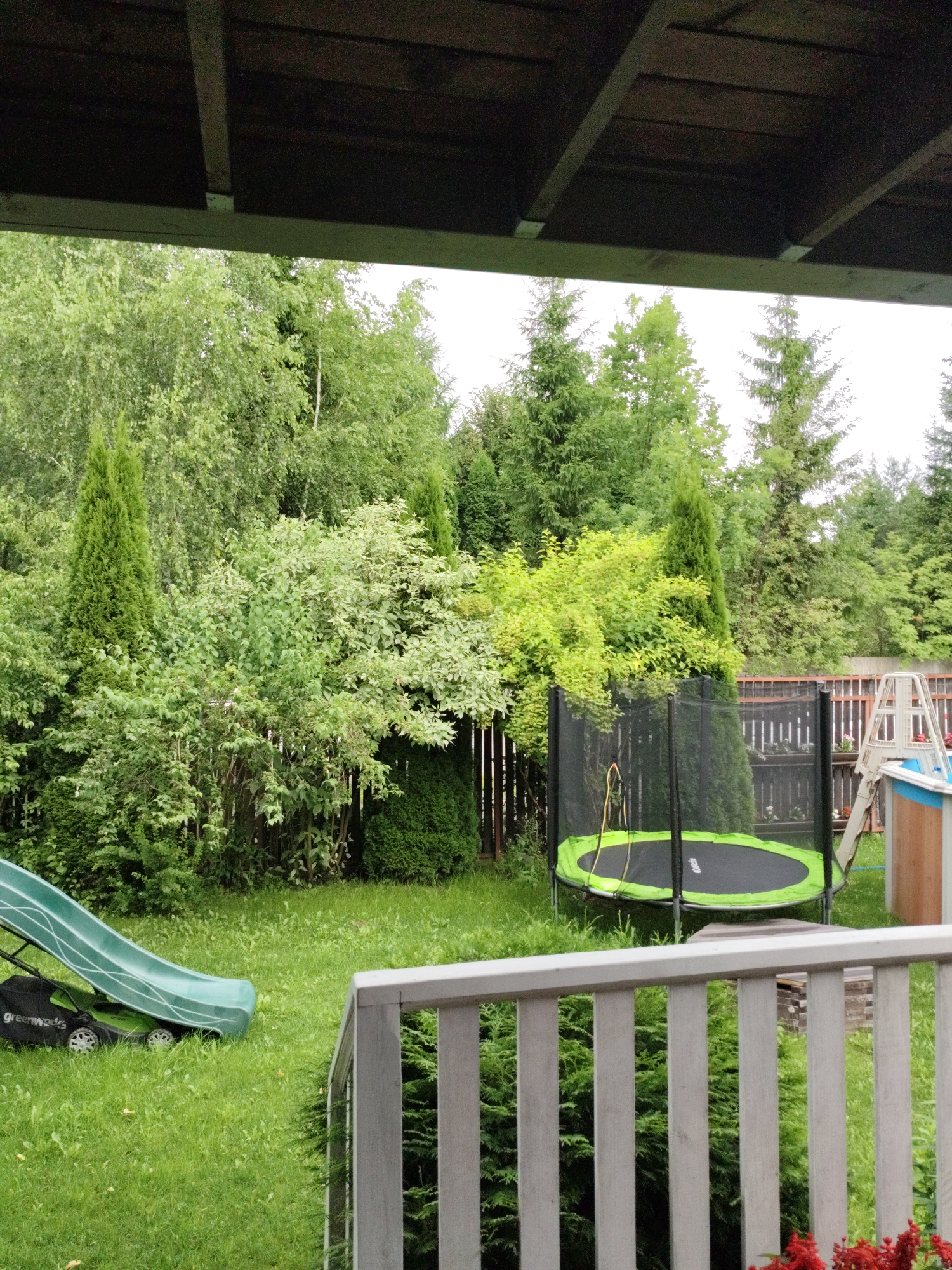 The image shows a backyard in Ермолино-2 Снт, Russia. The scene is dominated by lush green grass and a variety of trees and bushes. In the foreground, a wooden railing is visible, suggesting a porch or deck. To the left, a green children's slide is positioned over a green lawnmower. Further into the yard, a black trampoline with a neon green mat and enclosure is set up. Behind the trampoline, a fence and part of a swimming pool structure are visible. The overall environment is that of a well-maintained suburban backyard, likely used for recreation. The sky is overcast, indicating a cloudy day.