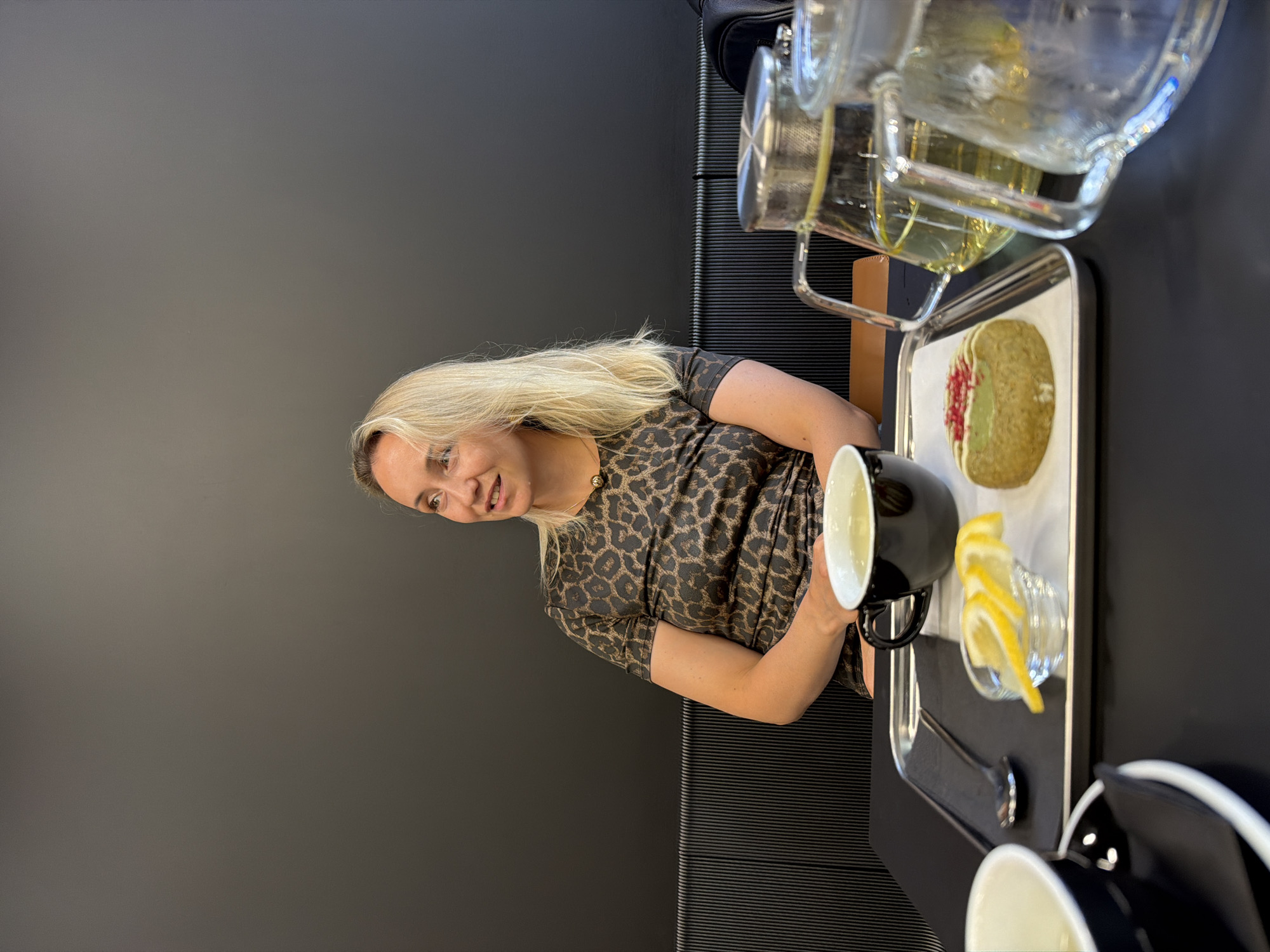 A blonde woman with fair skin and light makeup is sitting at a table in what appears to be a cafe. She is wearing a short-sleeved, brown and black leopard-print dress and a gold necklace with a circular pendant. Her hair is shoulder-length and blonde. She is looking to her right, with a slight smile.
On the table in front of her is a silver tray. On the tray, there is a black teacup filled with a light-colored liquid, a small glass bowl containing three lemon slices, and a small, round pastry topped with green frosting and red sprinkles. There is also a spoon on the tray. To the right of the tray are two clear glass teapots with metal infusers, both containing a yellow liquid. To the left of the tray, partially visible, is another white teacup.
The background consists of a dark gray wall and a dark ribbed bench. The table appears to be black. The lighting is moderate, creating some shadows.
This image was taken in Izhevsk, Russia.