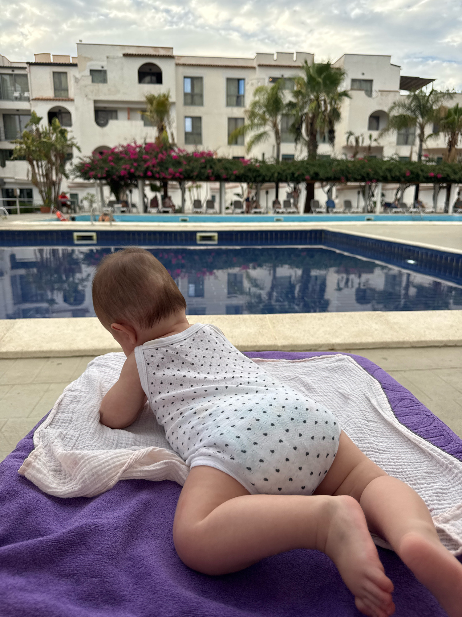 The image shows a baby lying face down on a purple towel and a white cloth on a sun lounger, with its back to the camera. The baby is wearing a white onesie with a pattern of small black hearts. In the background, there is a swimming pool with blue water and reflections of the surrounding buildings and palm trees. Beyond the pool, there are white buildings with balconies and arched windows, typical of Mediterranean architecture, and lush green trees and bushes. The sky is overcast. The scene appears to be at a resort or hotel complex. The location is identified as Isola delle Femmine, Italy.
