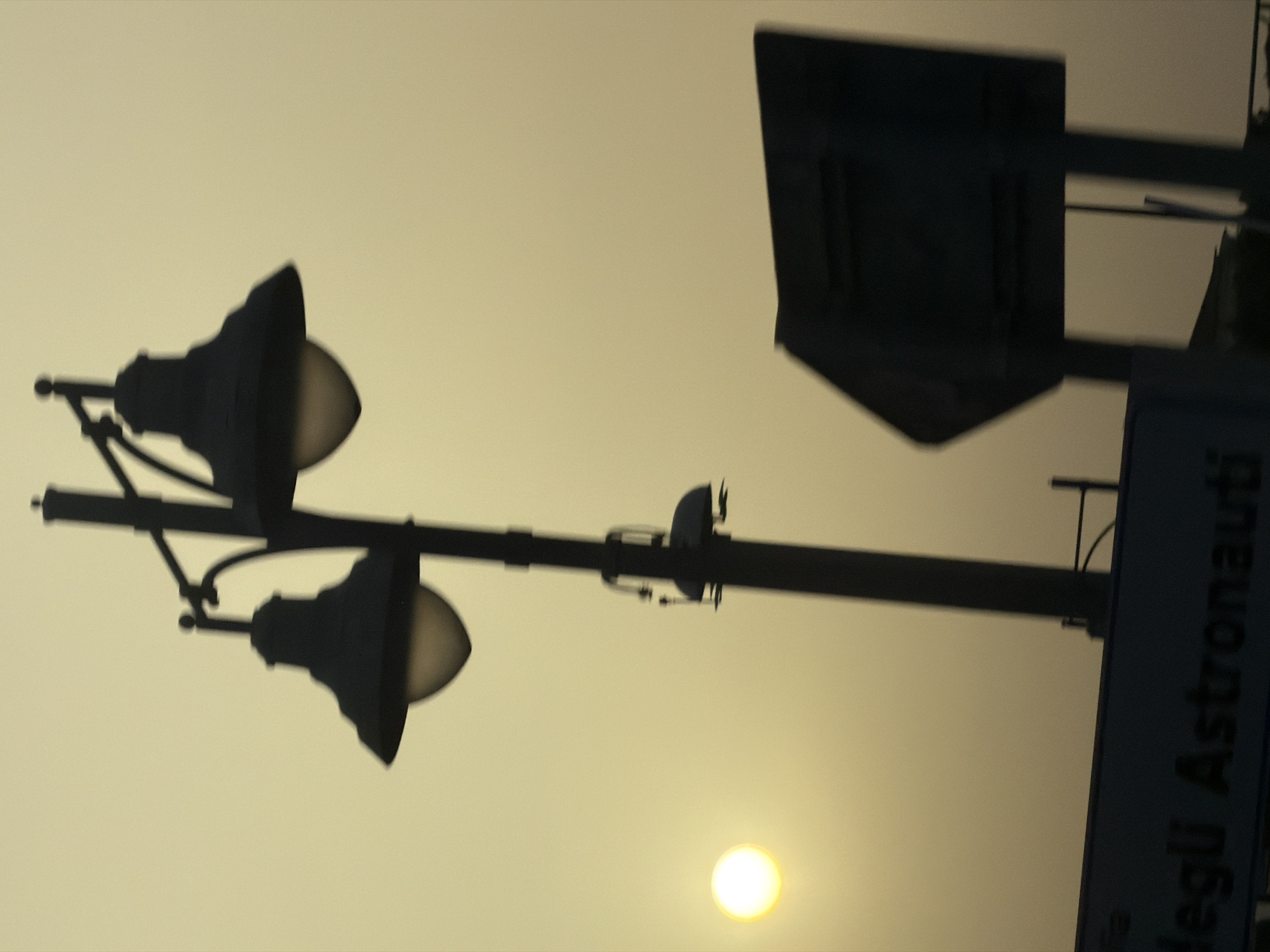 The image shows a street lamp with two lights and a sign in the city of Olbia, Italy. The sun is visible in the background, creating a hazy, golden-hour effect. The street lamp is silhouetted against the sky, with the two lights appearing as dark shapes with a faint glow. The sign, which is also silhouetted, has text on it, but it is difficult to read due to the lighting conditions. The overall impression is one of a serene and peaceful evening in Olbia.